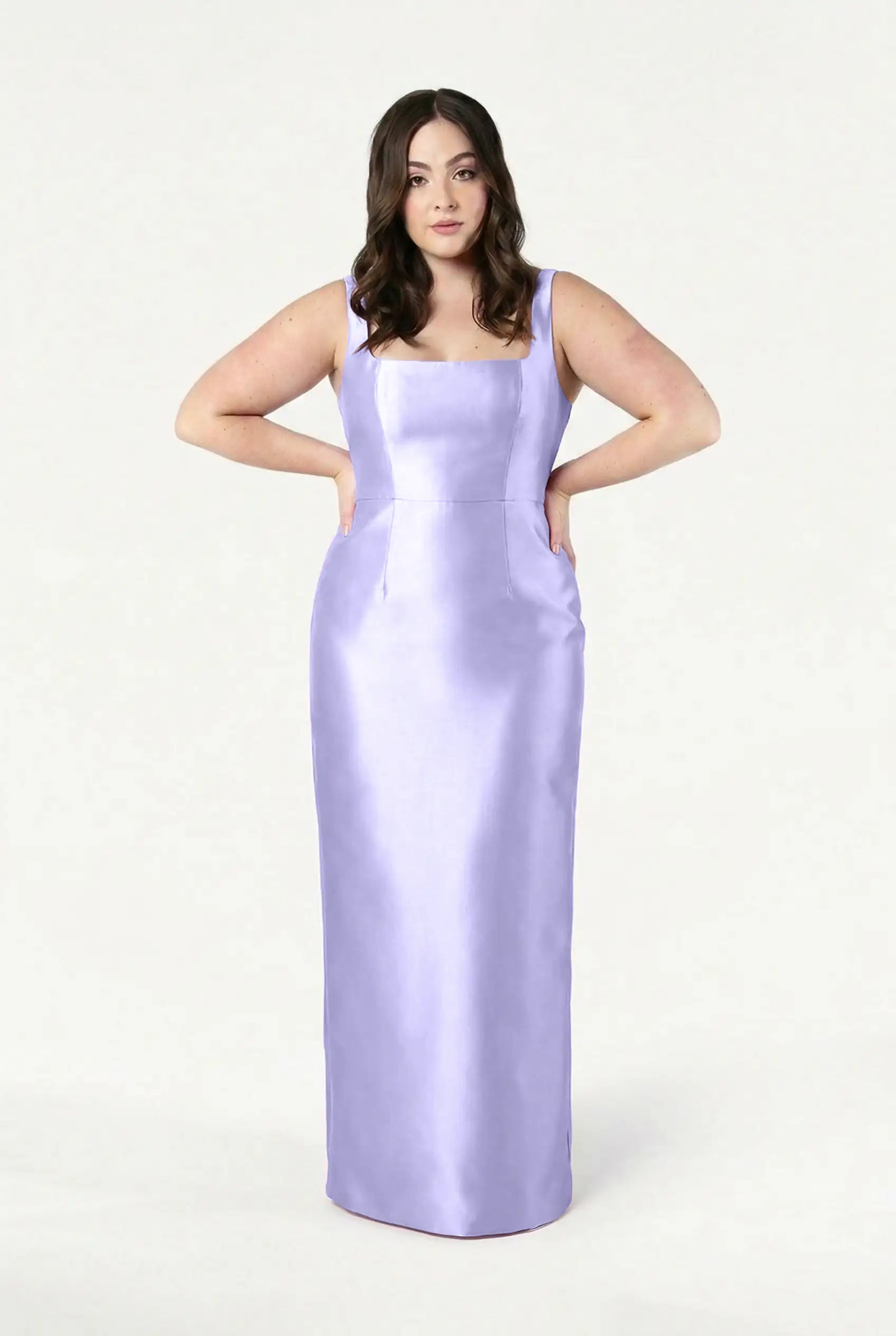 A woman with long brown hair stands with her hands on her hips, modeling the TH&TH Savoy Bridesmaid Dress in Lilac Mist Satin Twill, a sleeveless floor-length gown, against a plain white background.