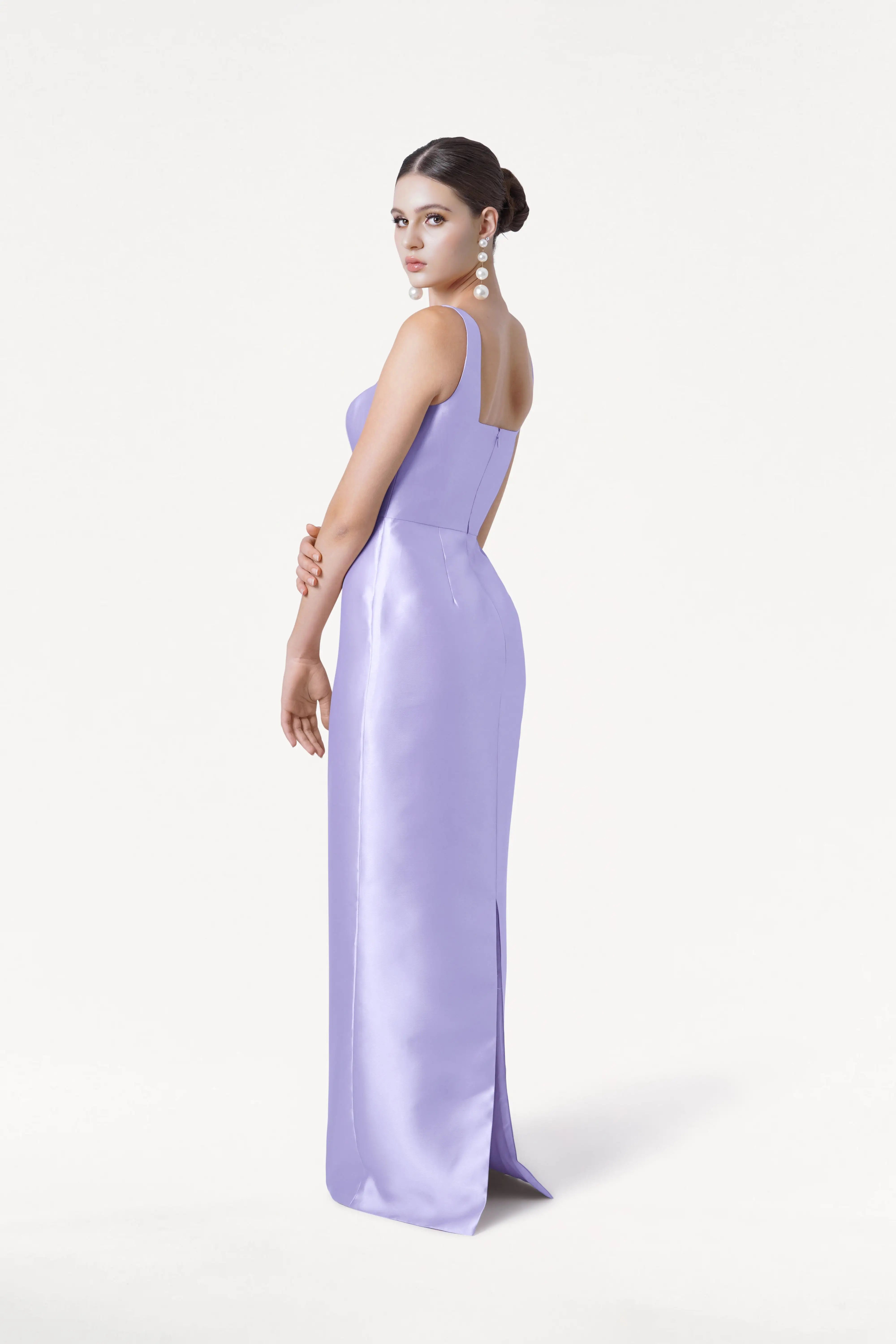 A woman with dark hair in an updo, wearing long pearl earrings and the TH&TH Savoy Bridesmaid Dress in Lilac Mist Satin Twill, stands looking over her left shoulder against a plain white background.