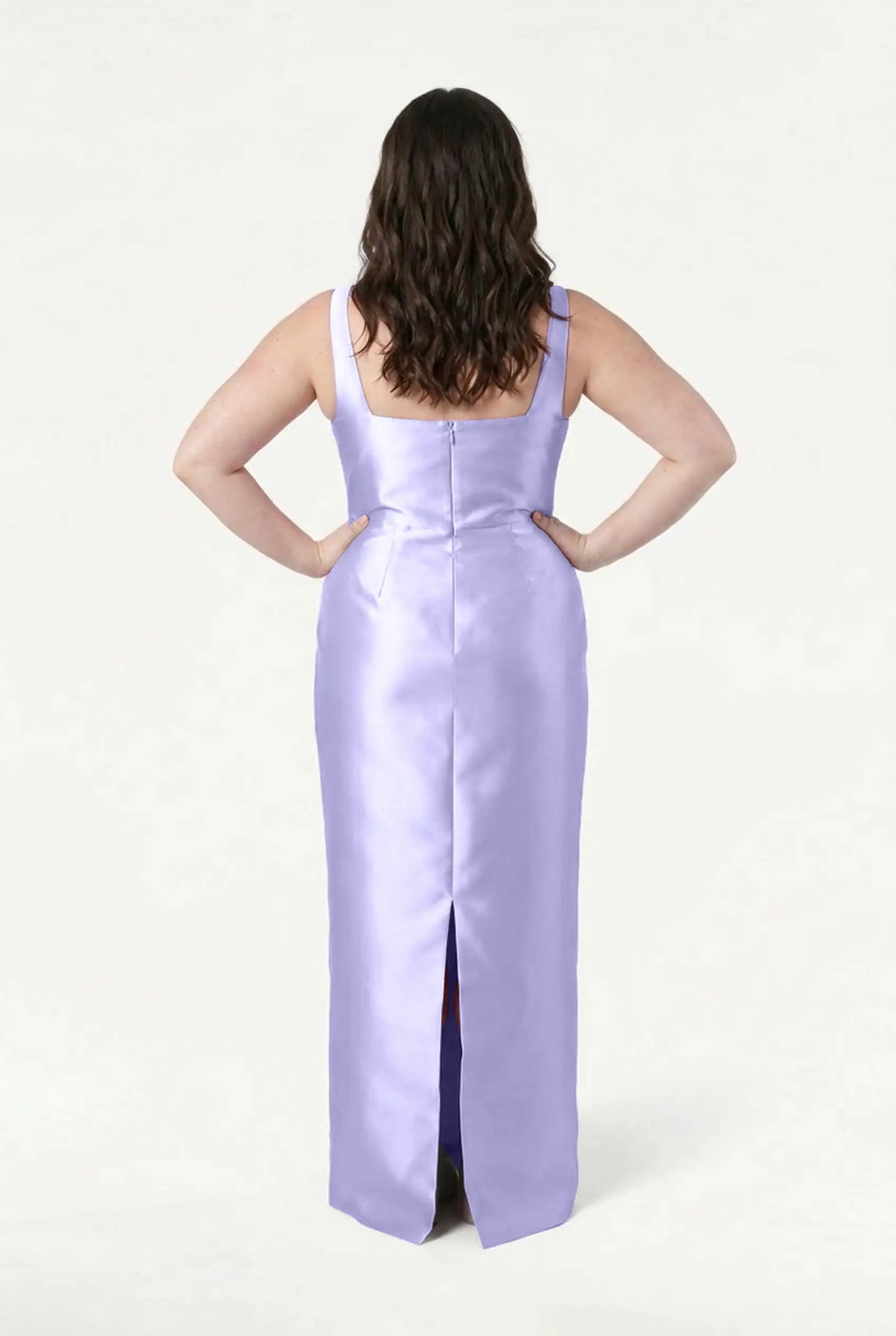 A woman with wavy brown hair stands with hands on hips, back facing, wearing the TH&TH Savoy Bridesmaid Dress in Lilac Mist Satin Twill—a long, sleeveless dress with a back slit—against a plain white background.