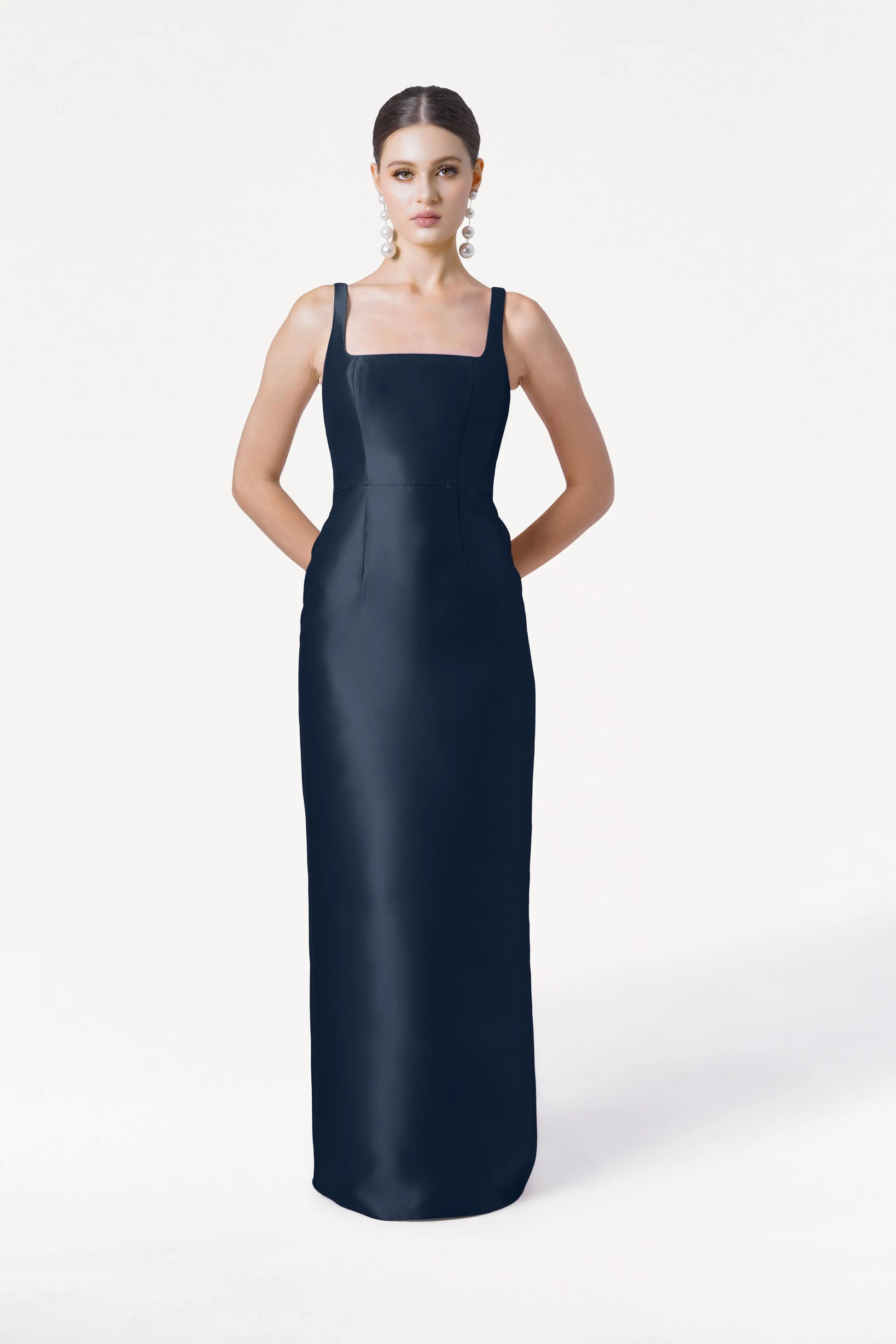 A woman with her hair pulled back and wearing large earrings poses against a white background in the TH&TH Savoy Bridesmaid Dress, a sleeveless, floor-length navy satin twill gown with a square neckline.