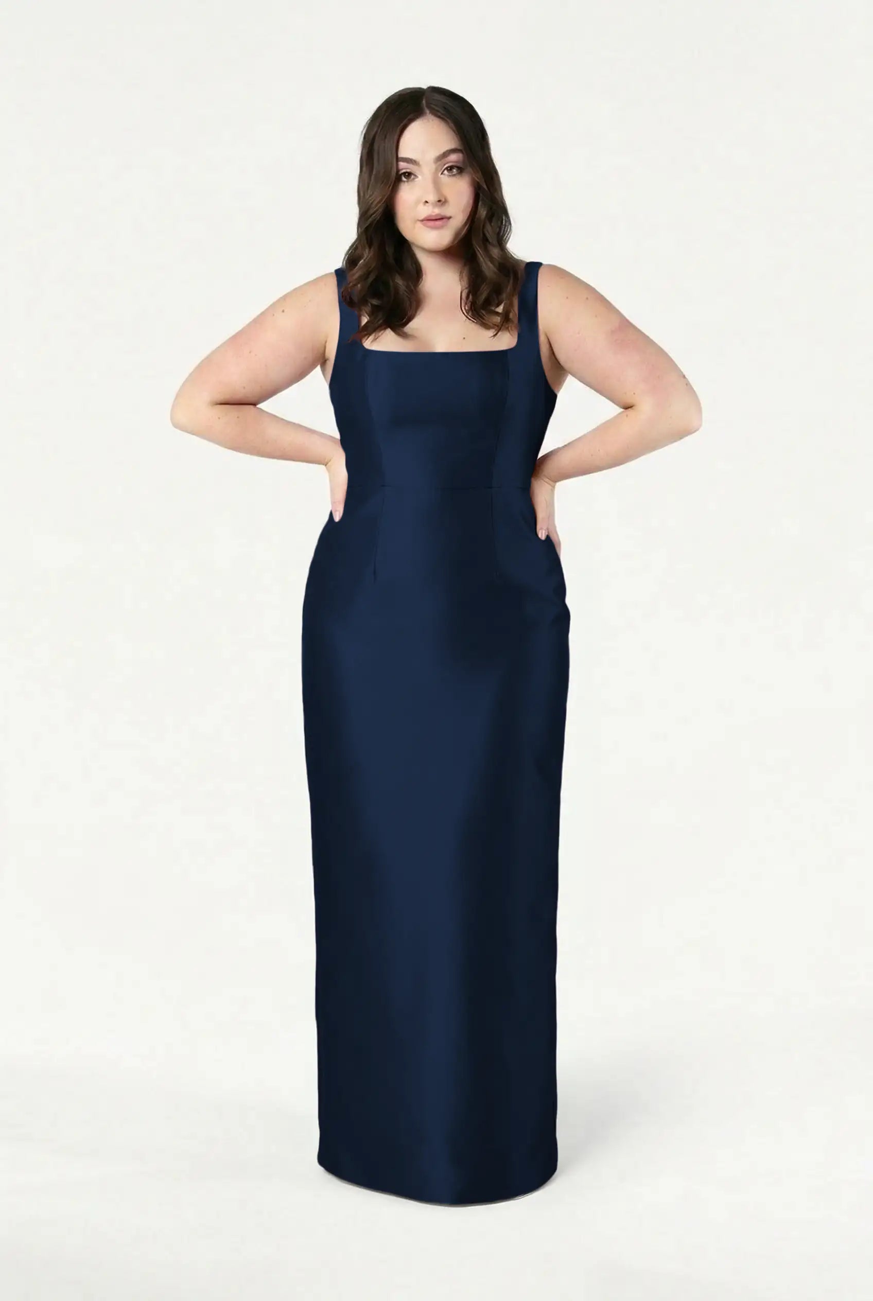 A woman with long brown hair stands against a white background, wearing the TH&TH Savoy Bridesmaid Dress in navy satin twill—a sleeveless, floor-length gown with a square neckline and her hands on her hips.