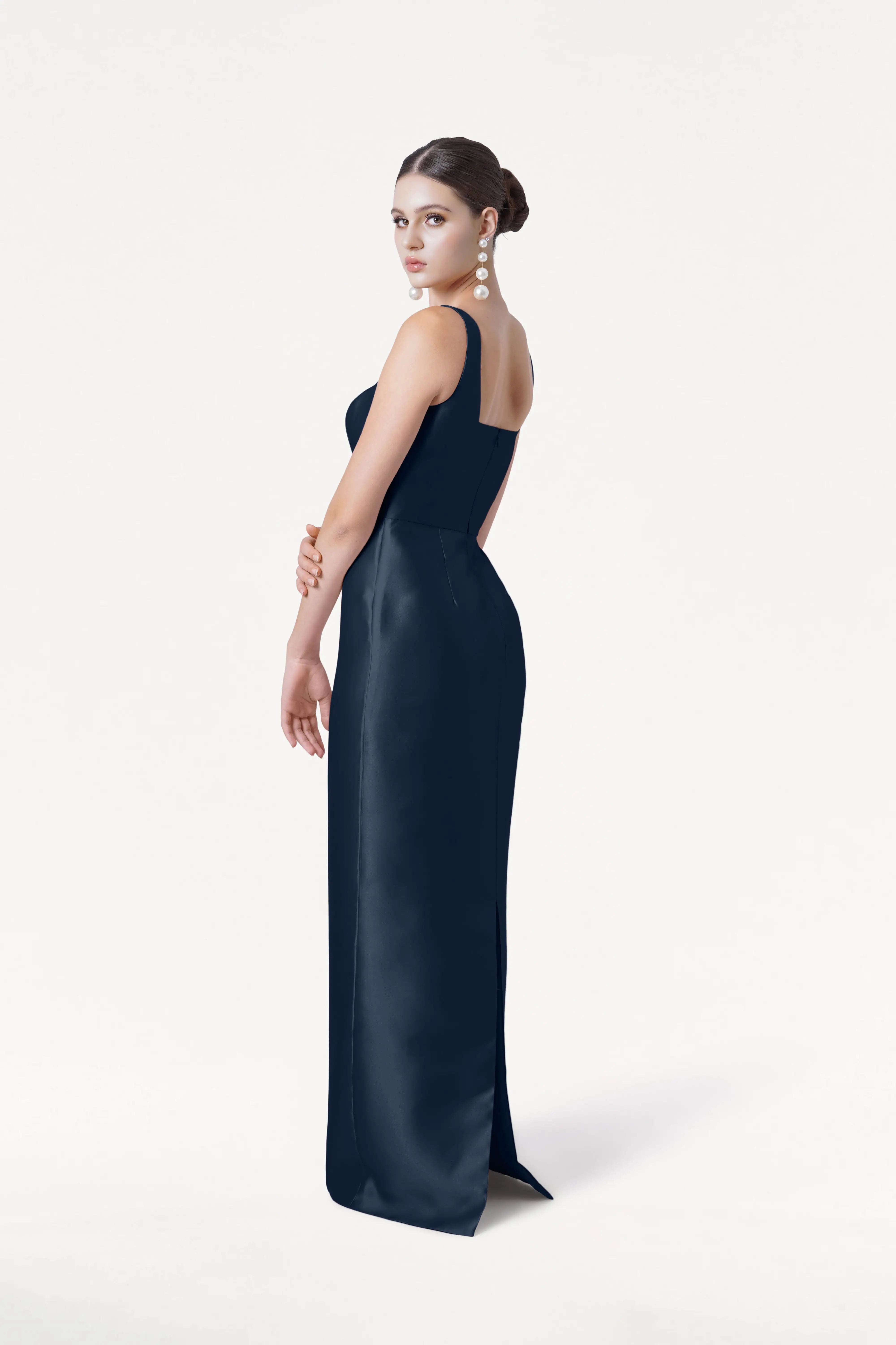 A woman with dark hair in a bun wears the TH&TH Savoy Bridesmaid Dress in Navy Satin Twill and large dangling pearl earrings, standing against a plain white background and looking over her shoulder.