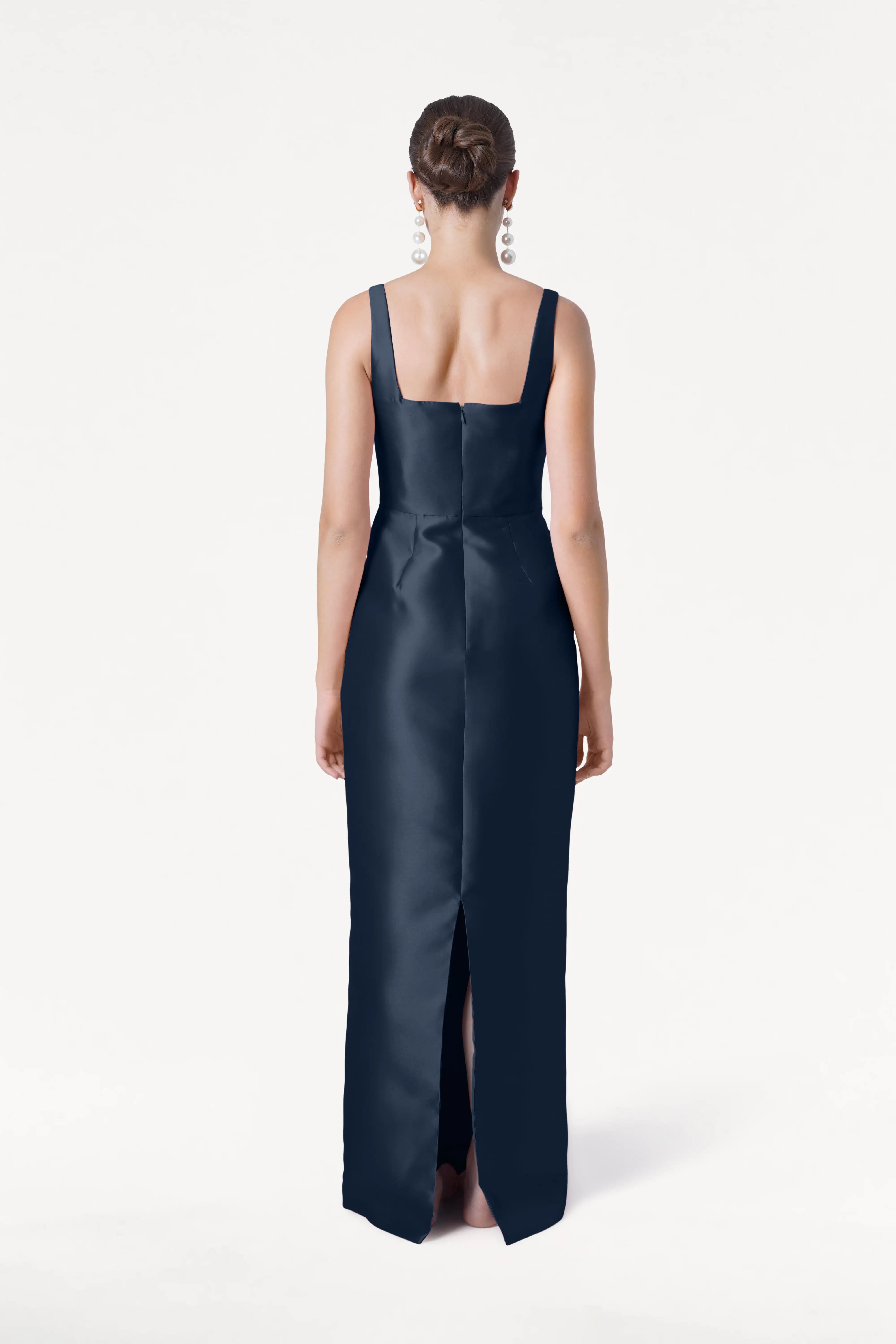 A woman with her hair in a bun and statement earrings faces away, wearing the TH&TH Savoy Bridesmaid Dress in Navy Satin Twill. This modern gown features a sleeveless design, square neckline, and back slit.