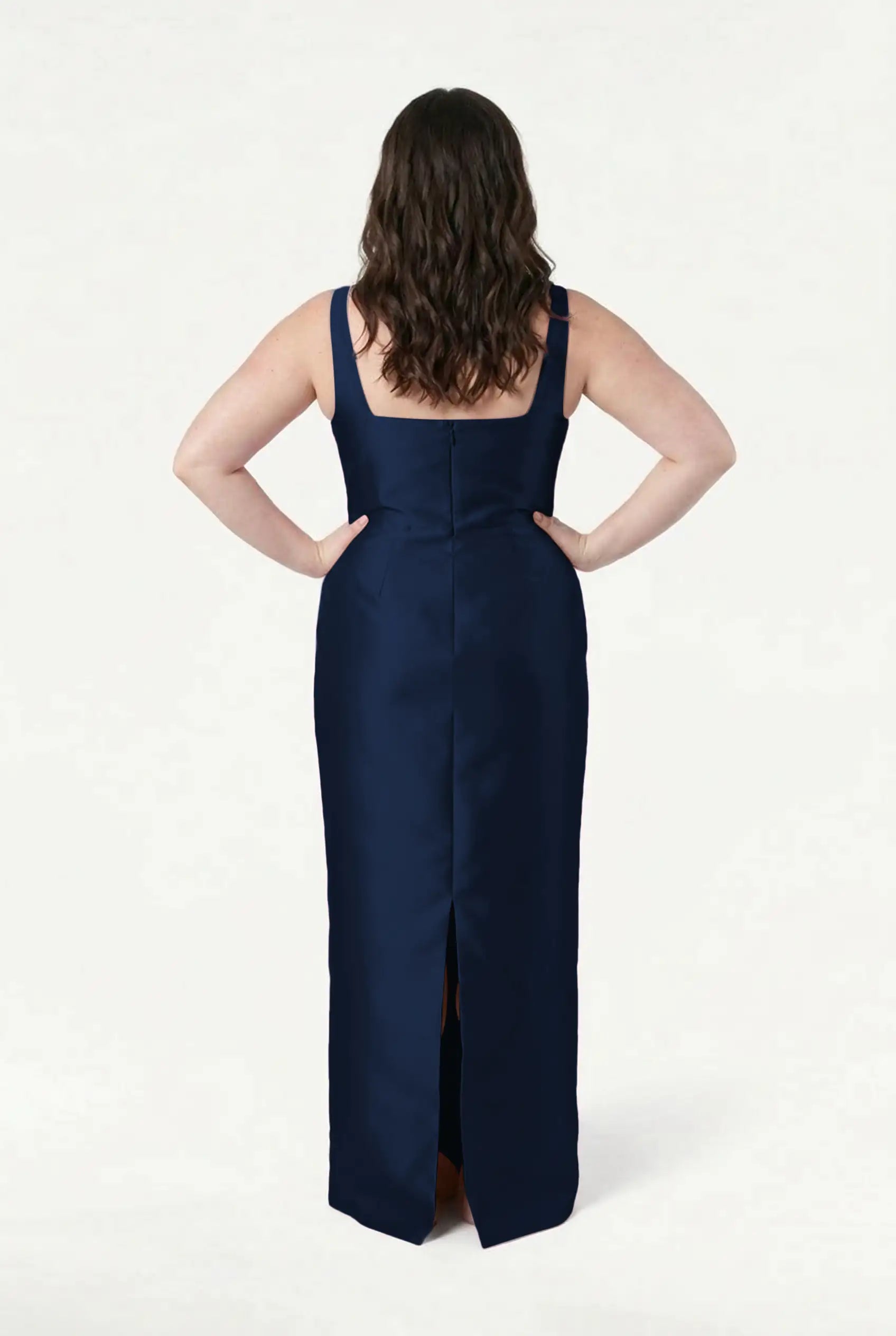 A woman with wavy brown hair stands with her hands on her hips, wearing the TH&TH Savoy Bridesmaid Dress in Navy Satin Twill—a sleeveless, floor-length gown with a back slit—set against a plain white background.
