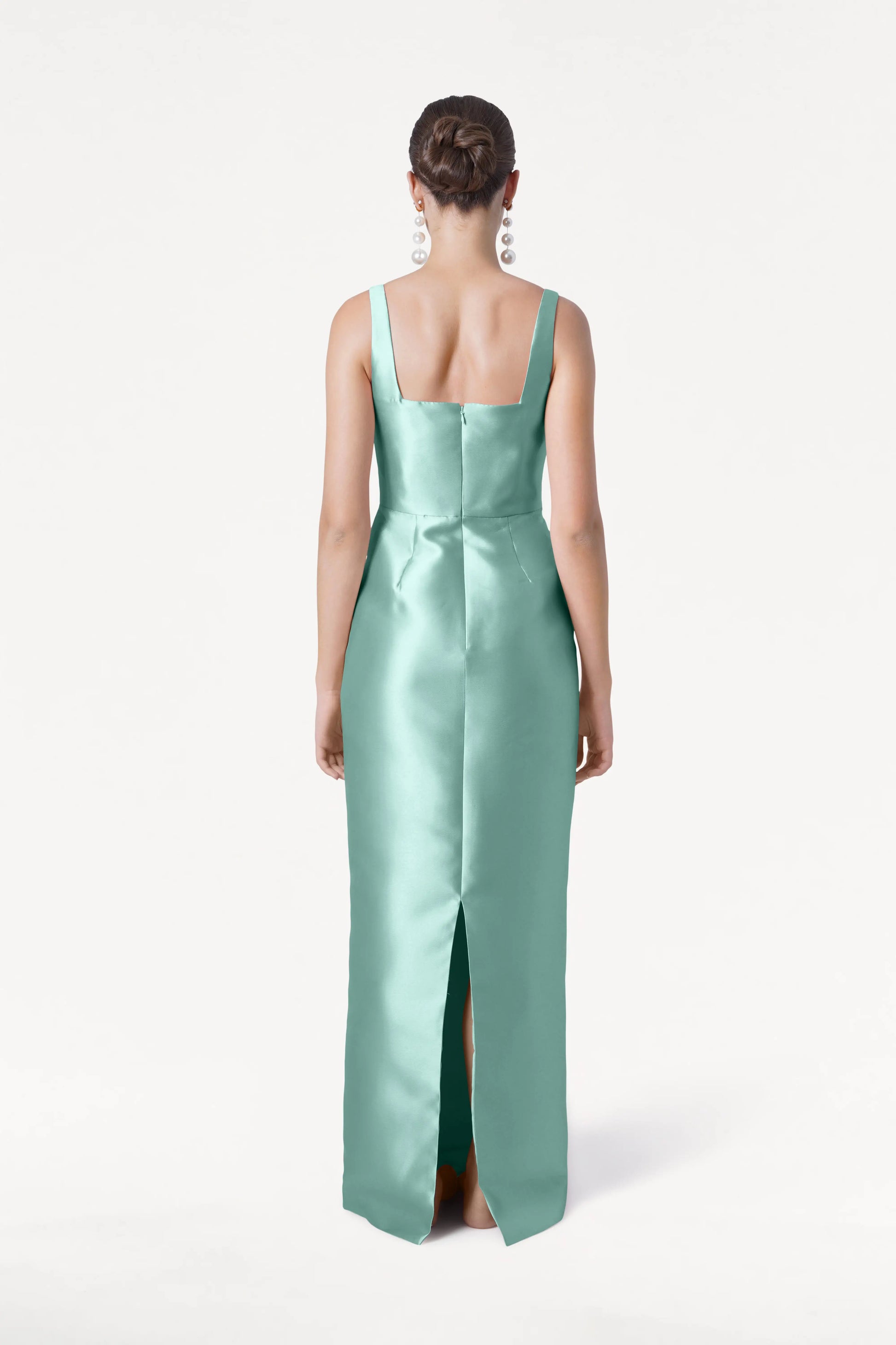A woman with her hair in a bun models the TH&TH Savoy Bridesmaid Dress in Seafoam Satin Twill, featuring a sleeveless design, modern square neckline, and back slit, against a plain white background.