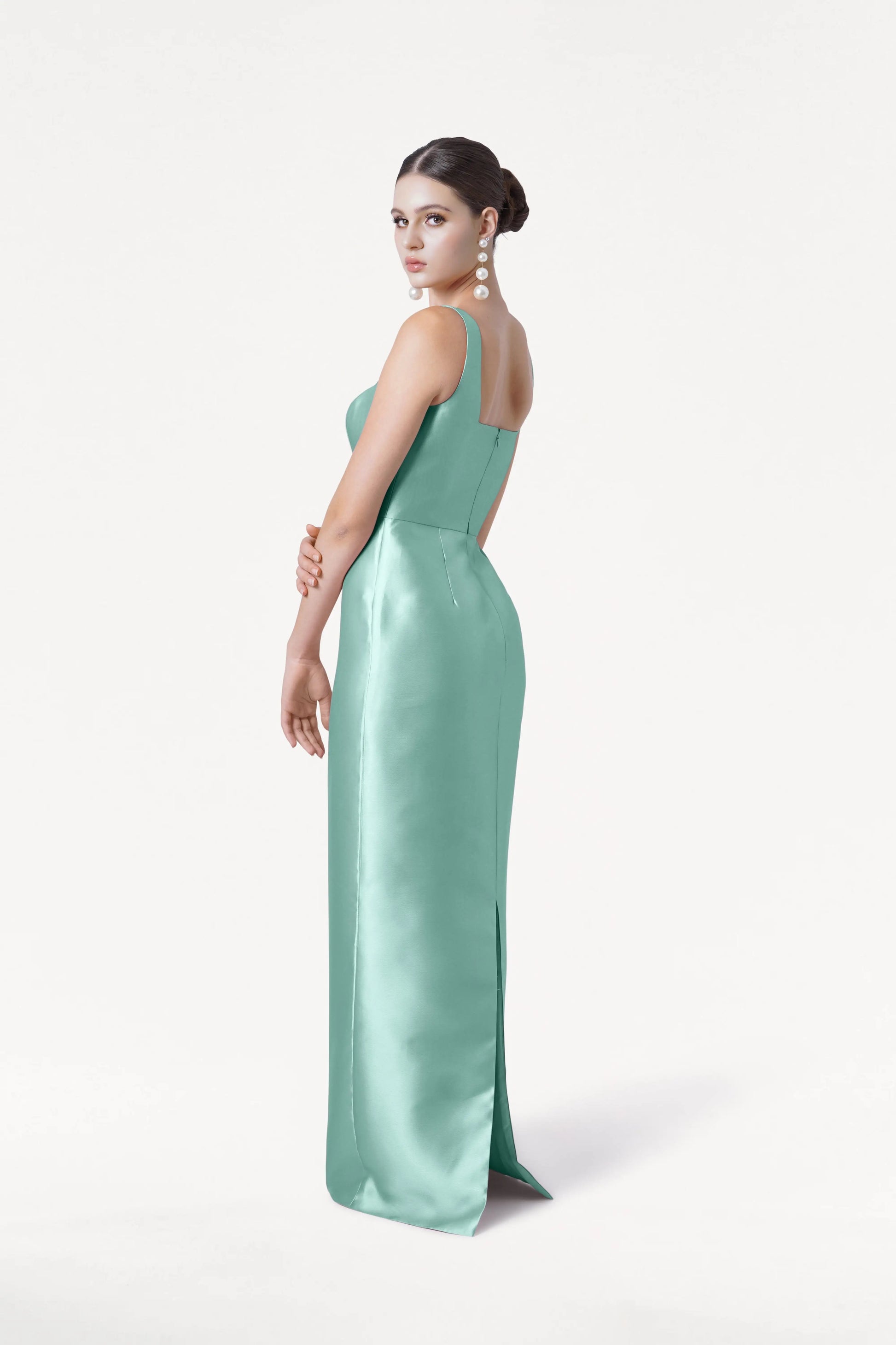 A woman wearing the TH&TH Savoy Bridesmaid Dress in Seafoam Satin Twill poses against a white background, her hair styled up, large pearl drop earrings on, glancing over her shoulder at the camera.