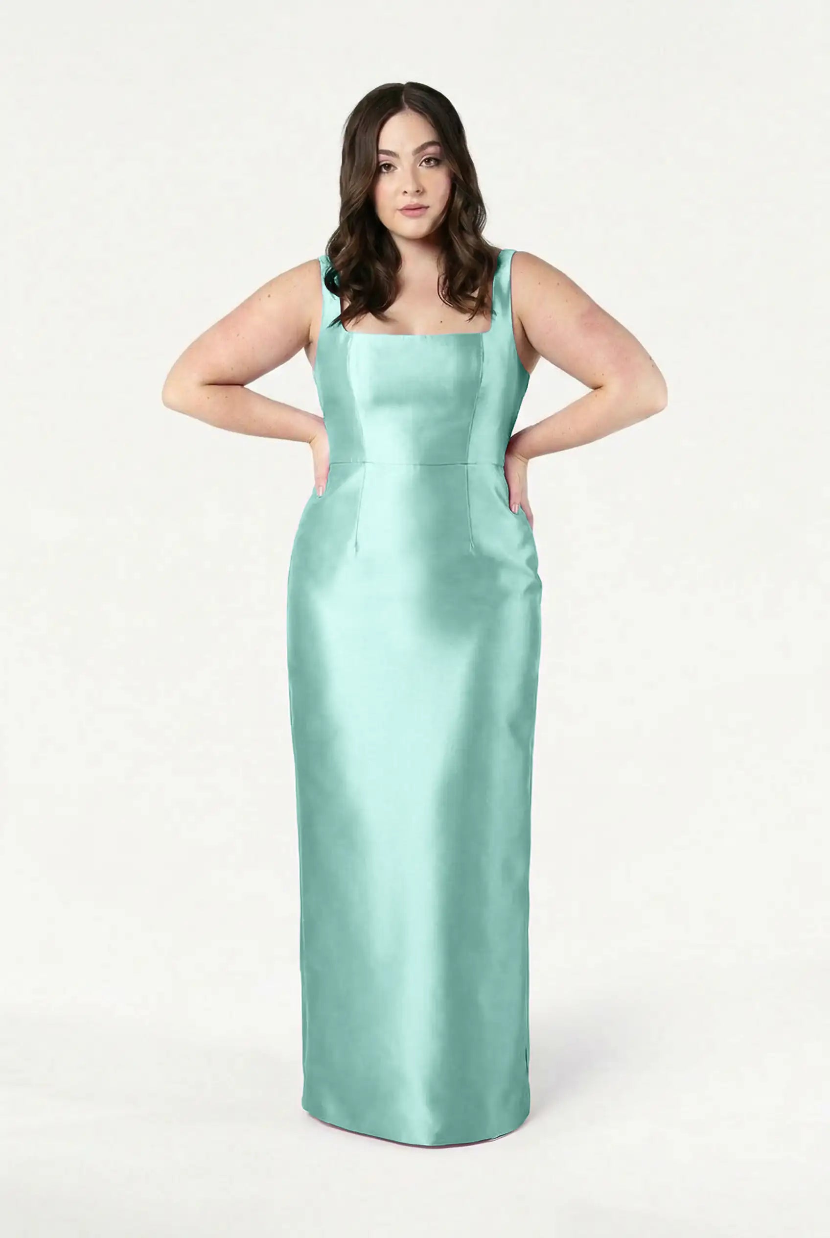 A woman with wavy brown hair wears the TH&TH Savoy Bridesmaid Dress in Seafoam Satin Twill, a sleeveless, floor-length gown with a modern square neckline, standing against a white background with her hands on her hips.
