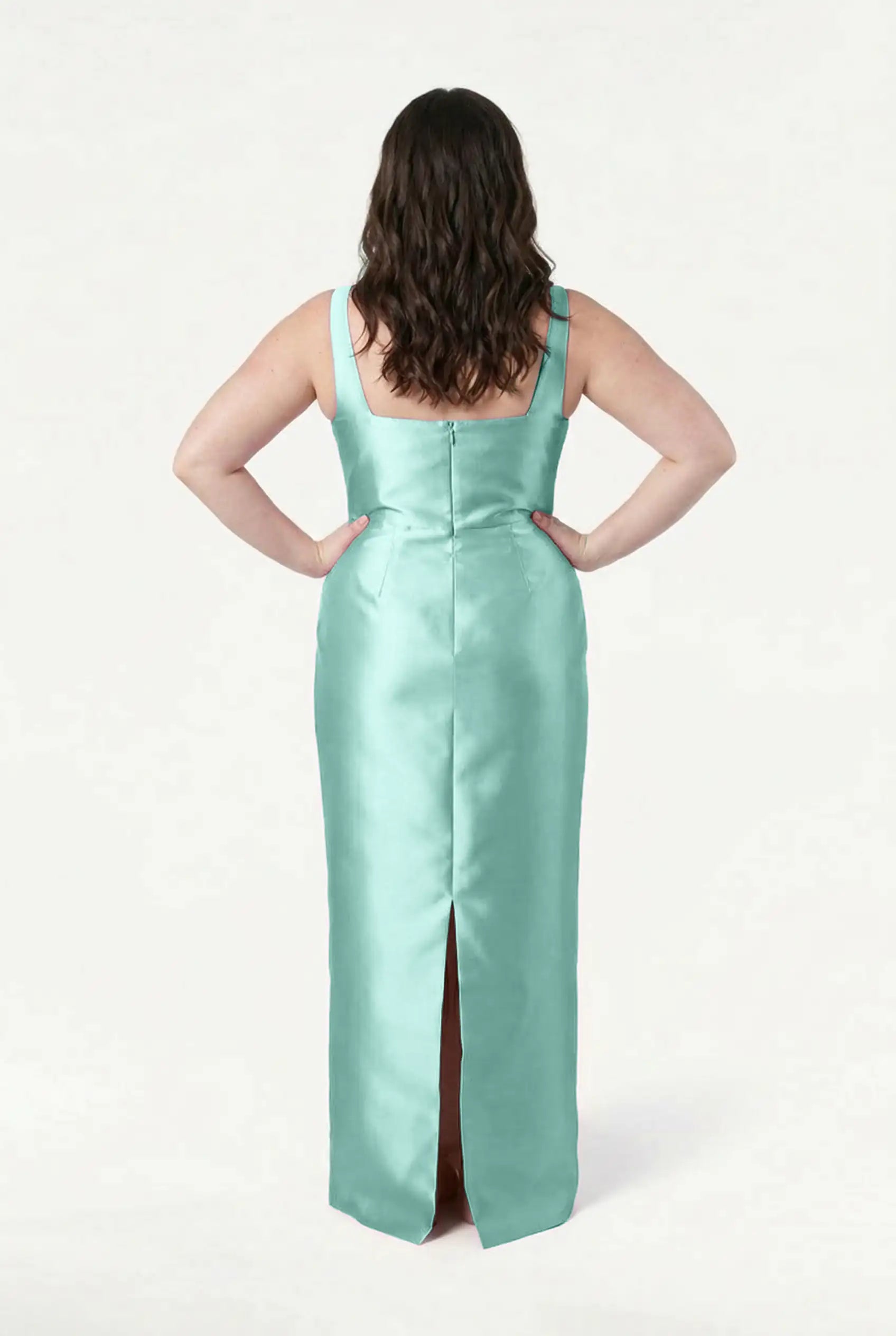 A woman with wavy brown hair stands with her back to the camera in a sleeveless TH&TH Savoy Bridesmaid Dress in Seafoam Satin Twill, featuring a modern square neckline and back slit, against a plain white background.