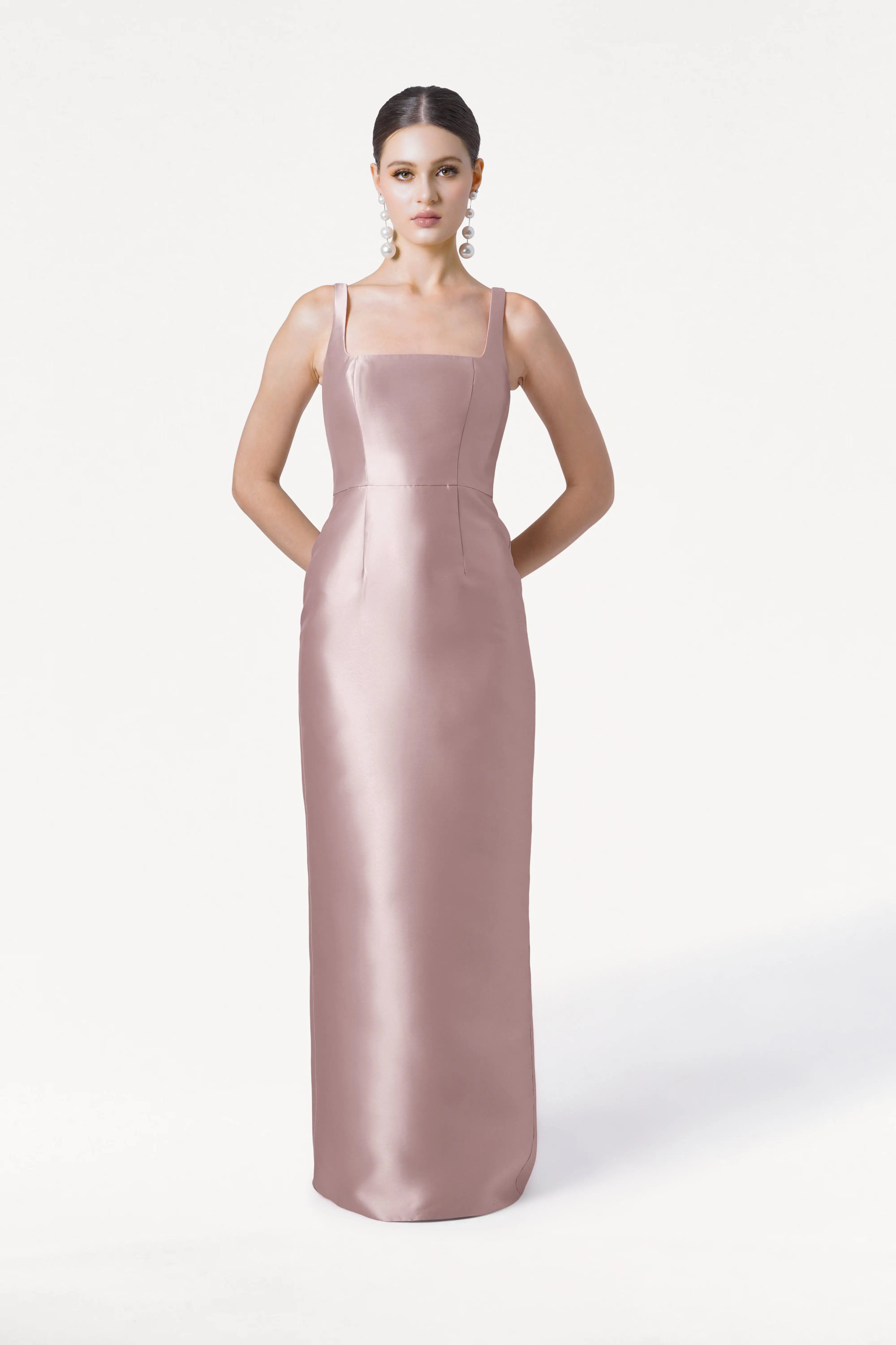 A woman poses with her hands behind her back, wearing the TH&TH Savoy Bridesmaid Dress in Smoked Blush Satin Twill, featuring a sleeveless, floor-length design and modern square neckline against a plain white background.