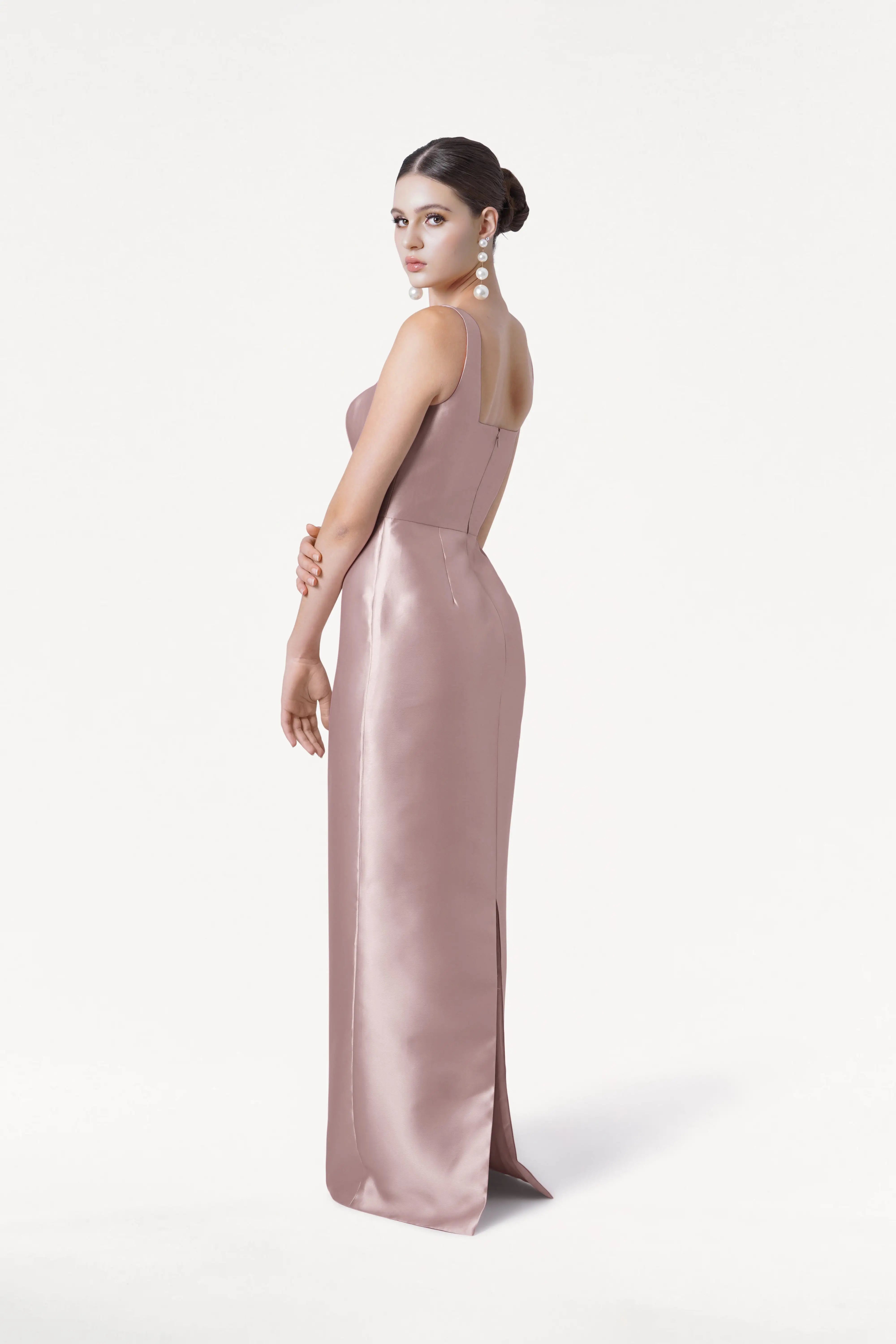 A woman models the TH&TH Savoy Bridesmaid Dress in Smoked Blush Satin Twill, posing sideways against a white backdrop with her hair in a bun and long pearl earrings.