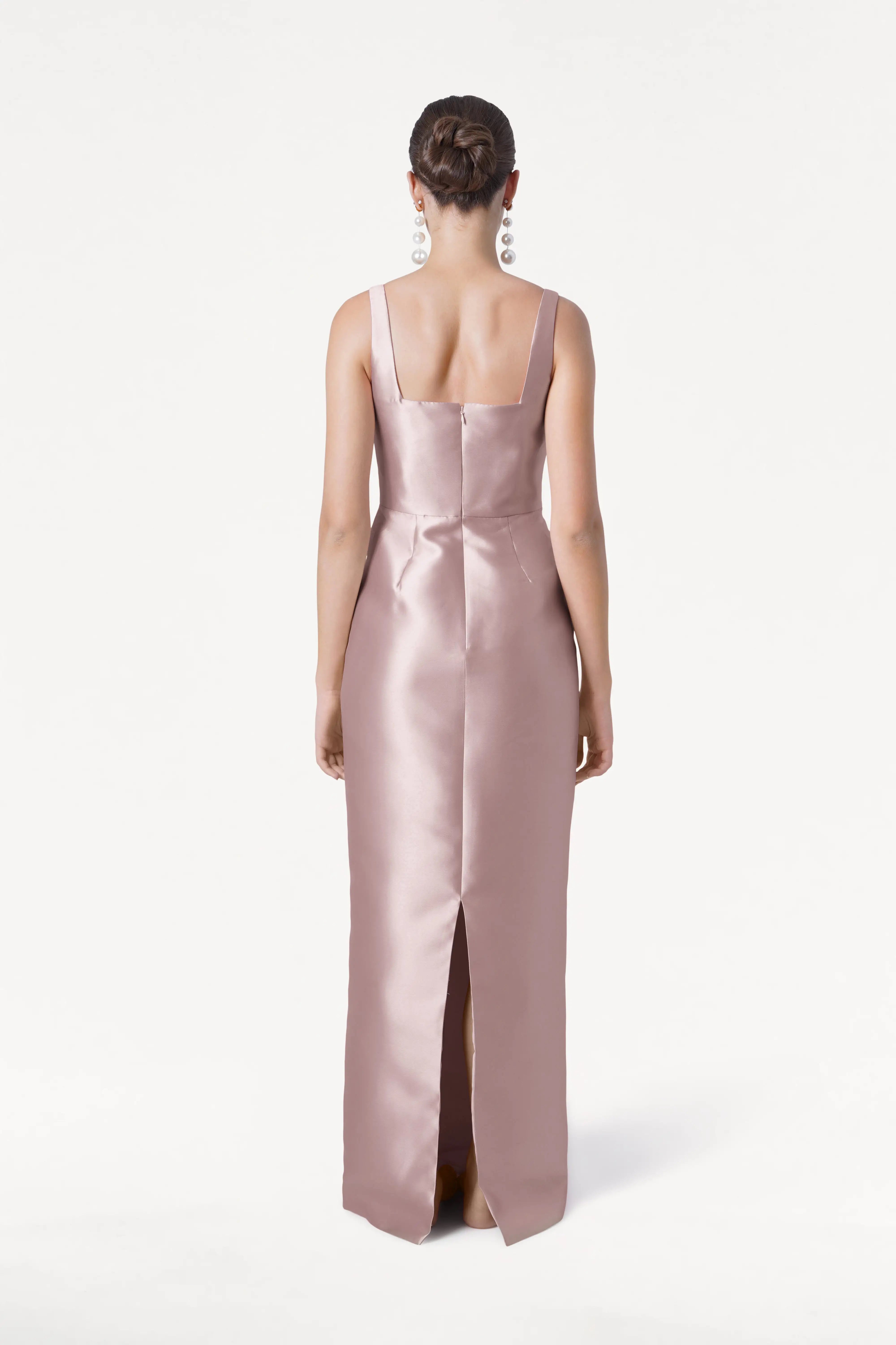 A woman faces away in the TH&TH Savoy Bridesmaid Dress, a sleeveless, floor-length gown in smoked blush satin twill with a modern square neckline and back slit, styled with her neat bun and statement earrings.