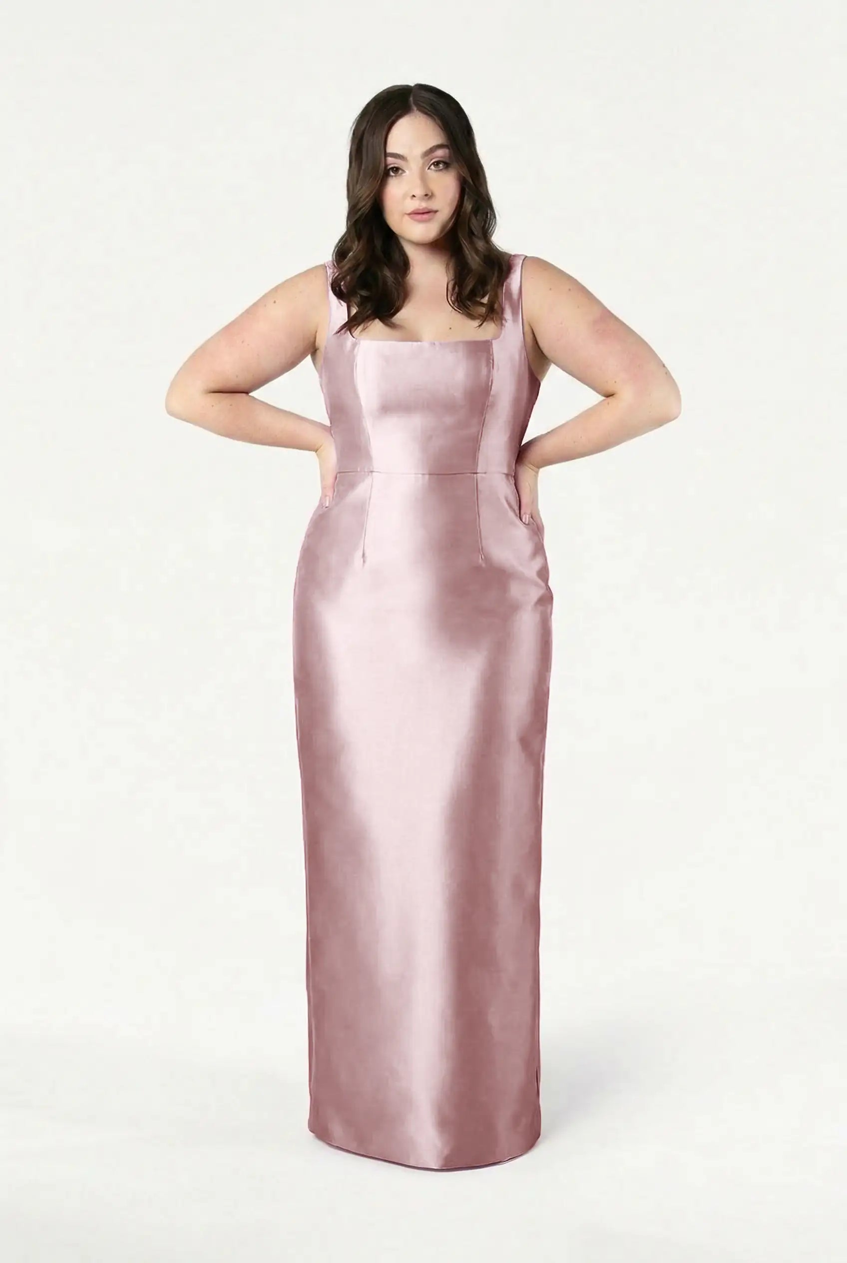 A woman with wavy brown hair models the TH&TH Savoy Bridesmaid Dress in Smoked Blush Satin Twill, a sleeveless, floor-length gown with a modern square neckline, standing hands on hips against a white background.