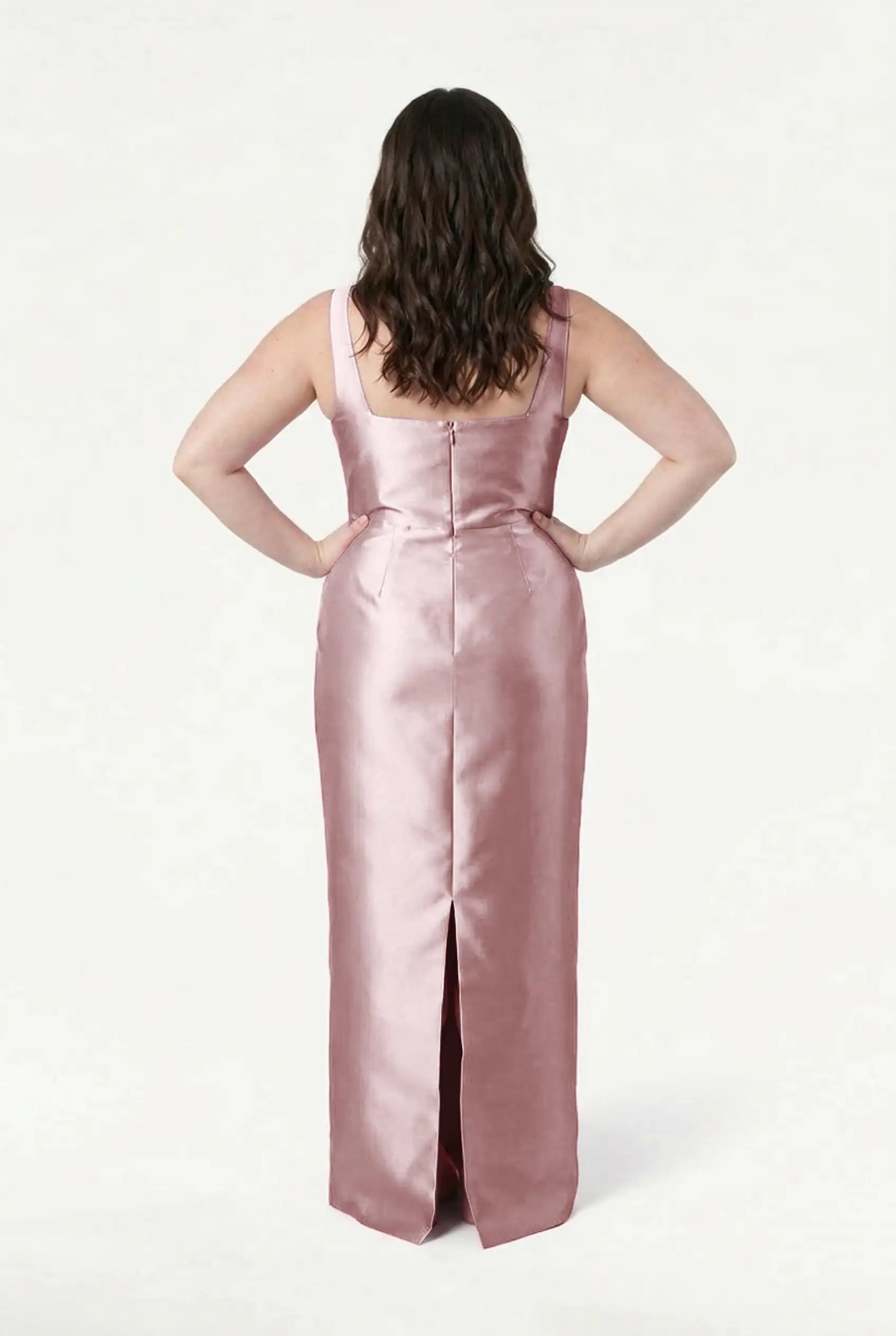 A woman with wavy brown hair stands hands on hips, modeling the TH&TH Savoy Bridesmaid Dress in Smoked Blush Satin Twill—sleeveless with a modern square neckline and back slit—photographed from behind against a plain white background.