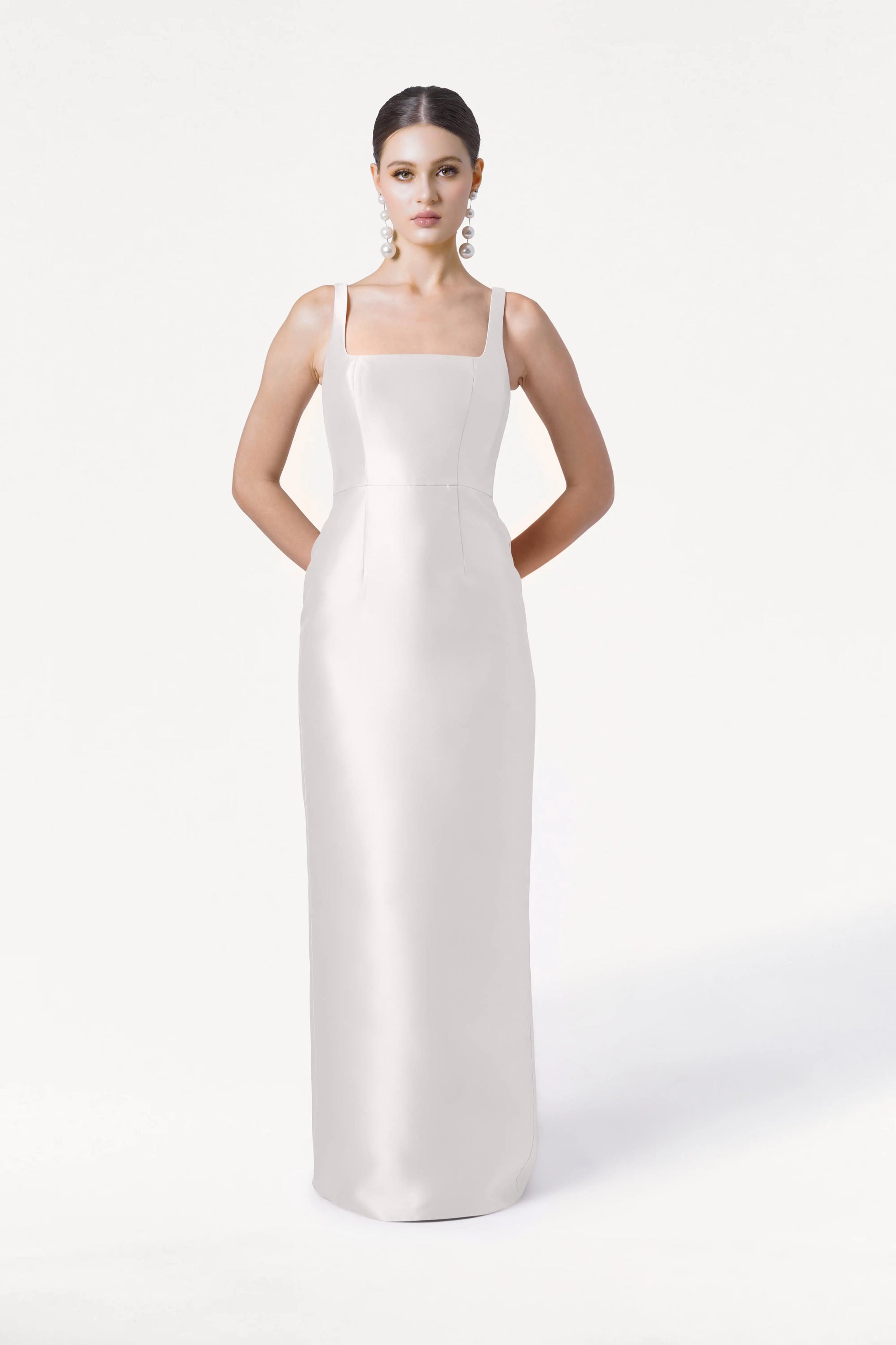 A woman poses against a white background wearing the Savoy Dress in Ivory Satin Twill by TH&TH, a sleek, floor-length bridesmaid dress with wide straps and a modern square neckline. She accessorizes with bold dangling earrings.