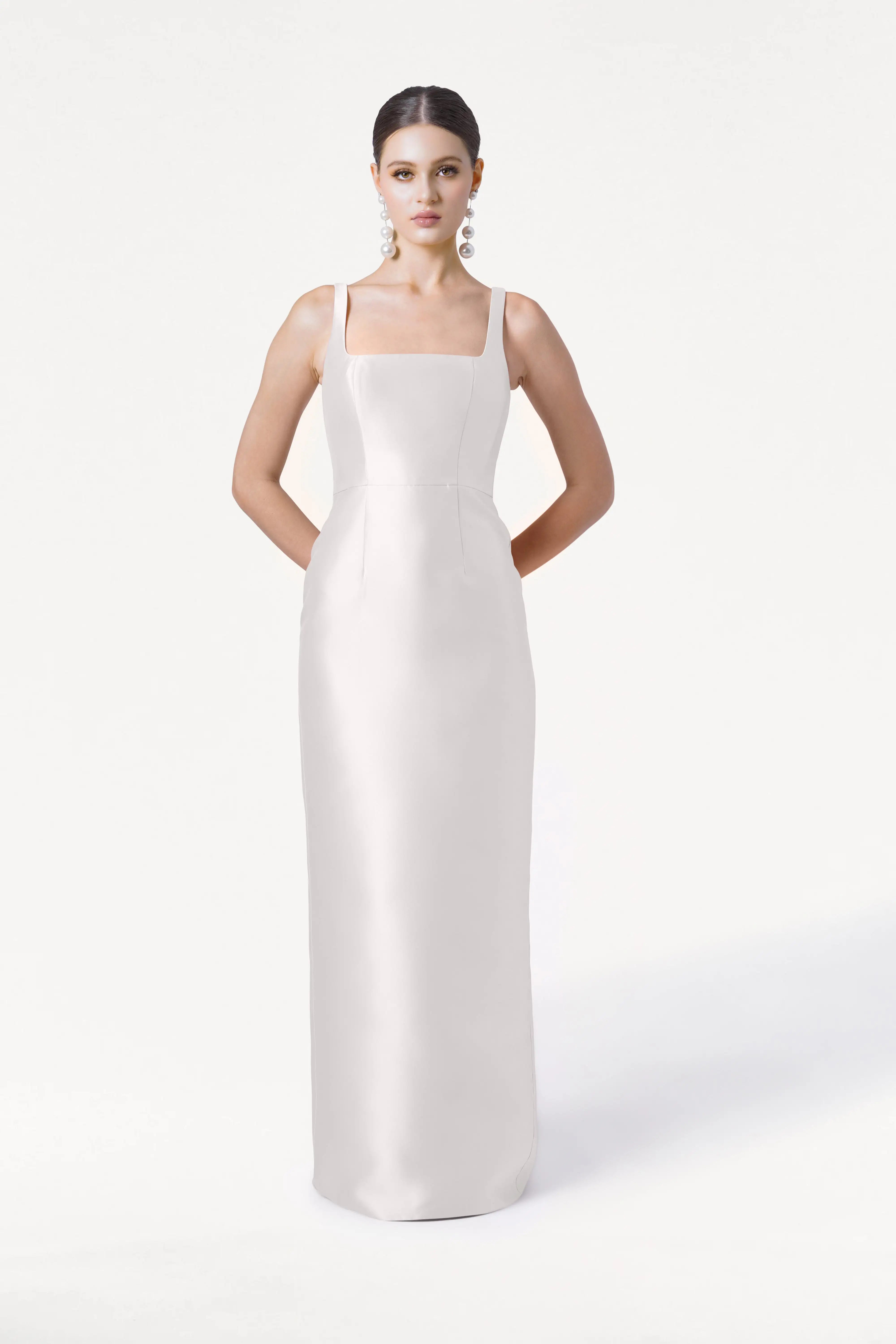 A woman poses against a white background wearing the Savoy Dress in Ivory Satin Twill by TH&TH, a sleek, floor-length bridesmaid dress with wide straps and a modern square neckline. She accessorizes with bold dangling earrings.