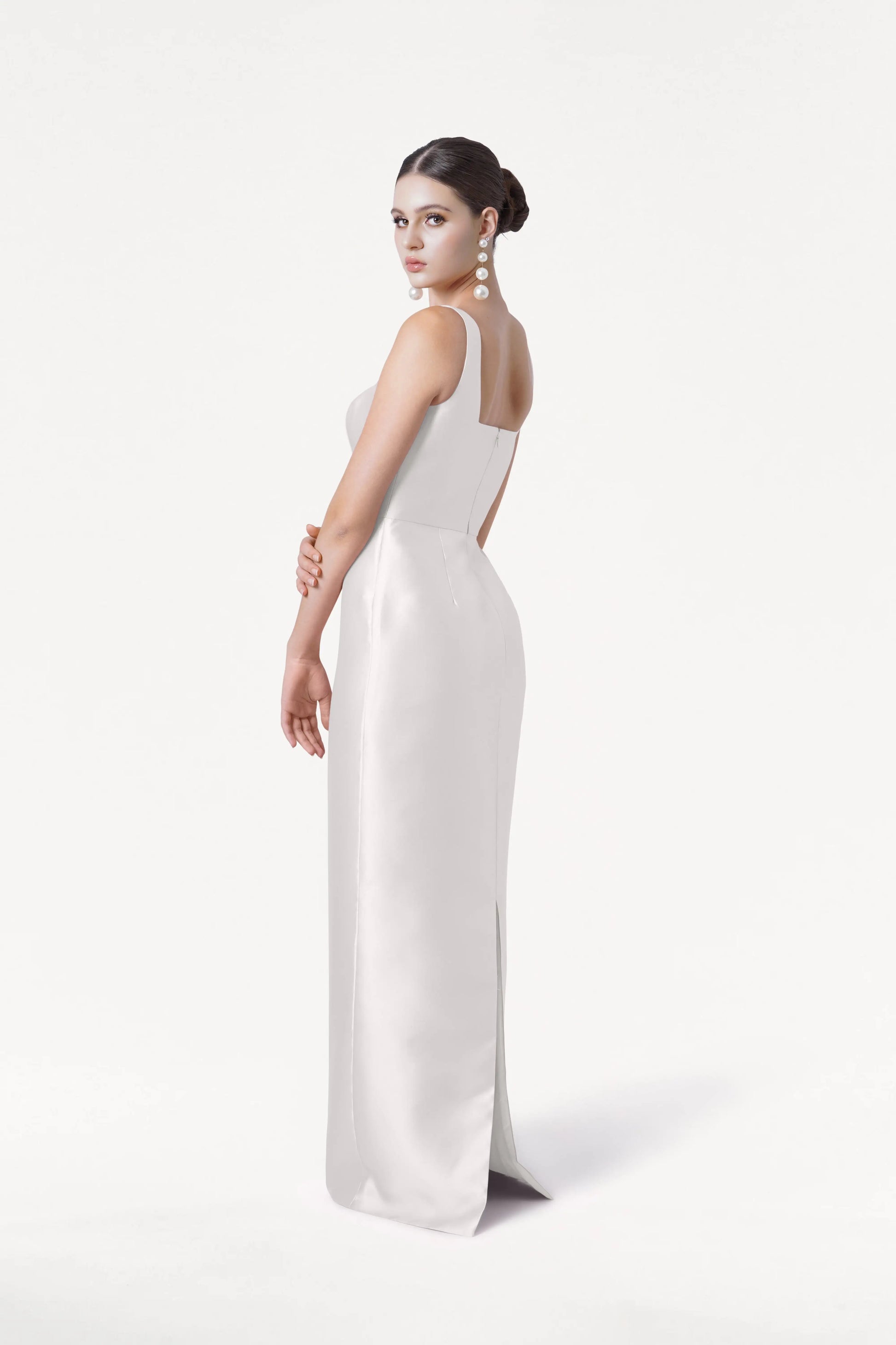 A woman wears the Savoy Dress in Ivory Satin Twill by TH&TH, featuring a modern square neckline. She stands against a white background, hair in a neat bun, large pearl earrings on, posing with one arm bent and looking over her shoulder.