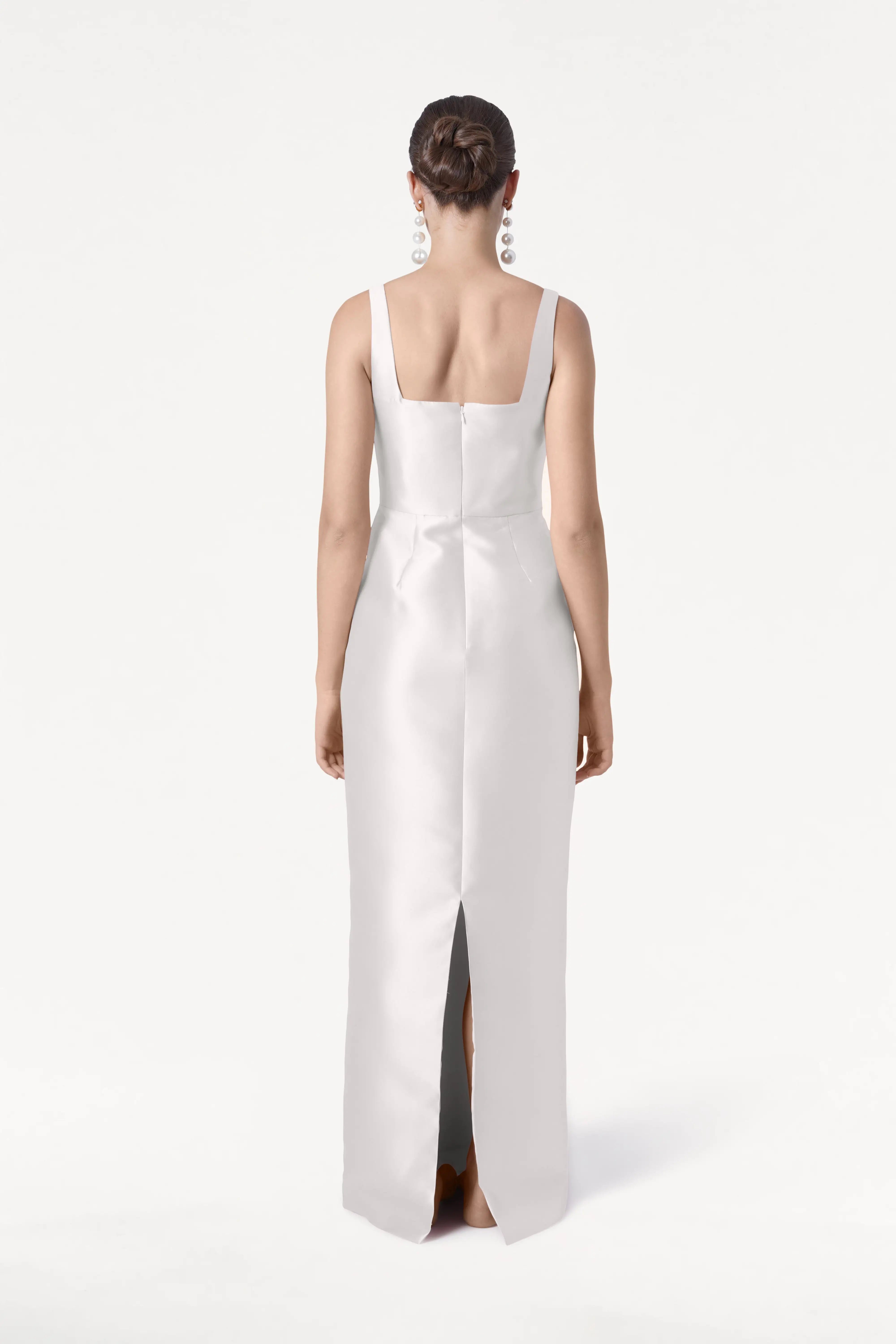 A woman stands facing away in the Savoy Dress by TH&TH, crafted from ivory satin twill. The gown has a modern square neckline, rear slit, and smooth finish. Her hair is styled in a neat bun and she wears large, dangling earrings.