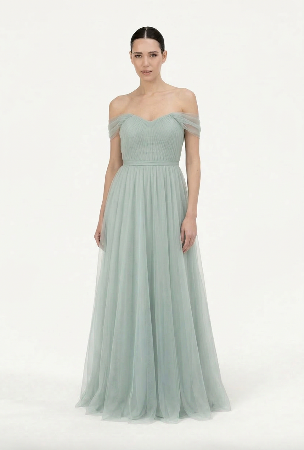 A woman wears the TH&TH Bardot Bridesmaid Dress in Dusty Eucalyptus Tulle, featuring an off-shoulder, fitted pleated bodice and a flowing tulle skirt, standing against a plain white background.