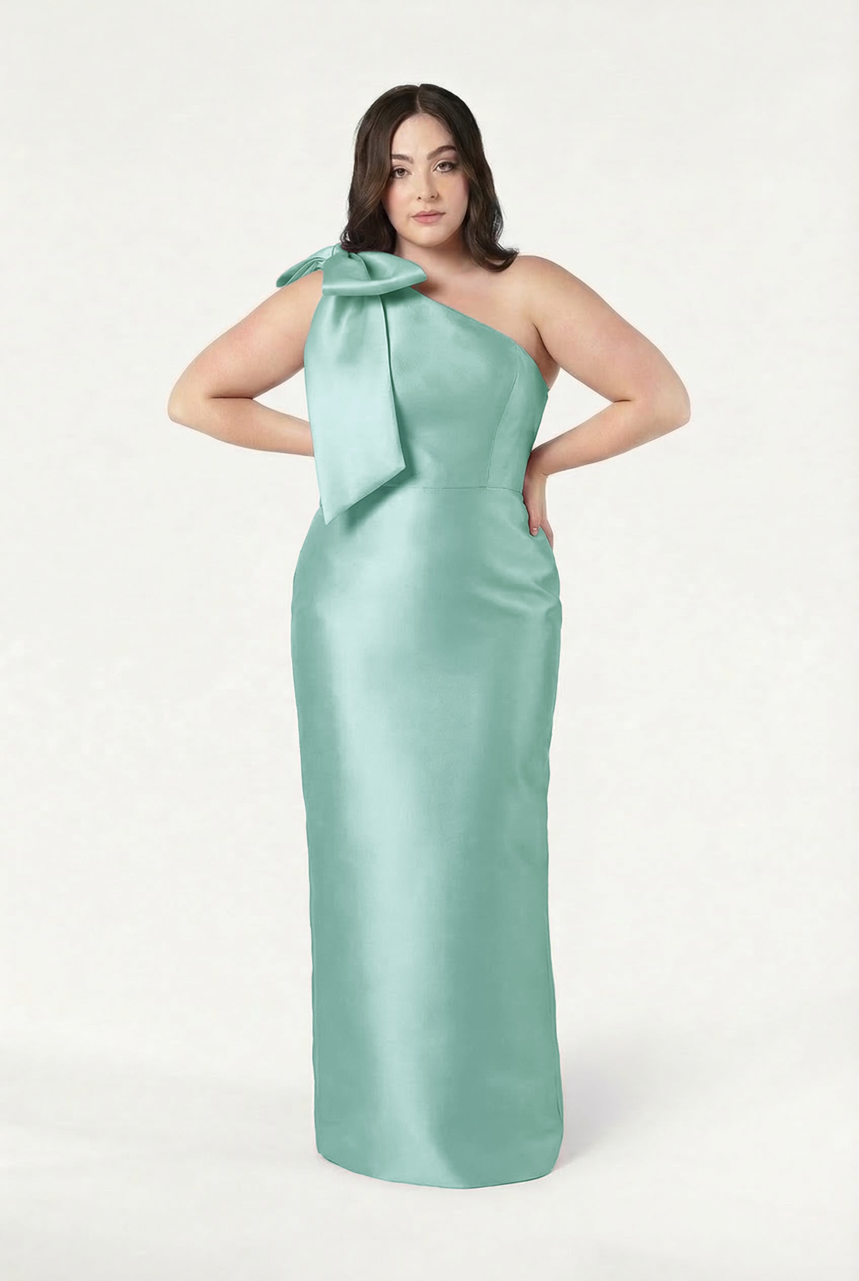 A woman wears the TH&TH Ritz Bridesmaid Dress in Seafoam Satin Twill, a sleeveless, floor-length gown with an asymmetrical one-shoulder neckline and a statement bow, posing with hands on her hips against a plain white background.