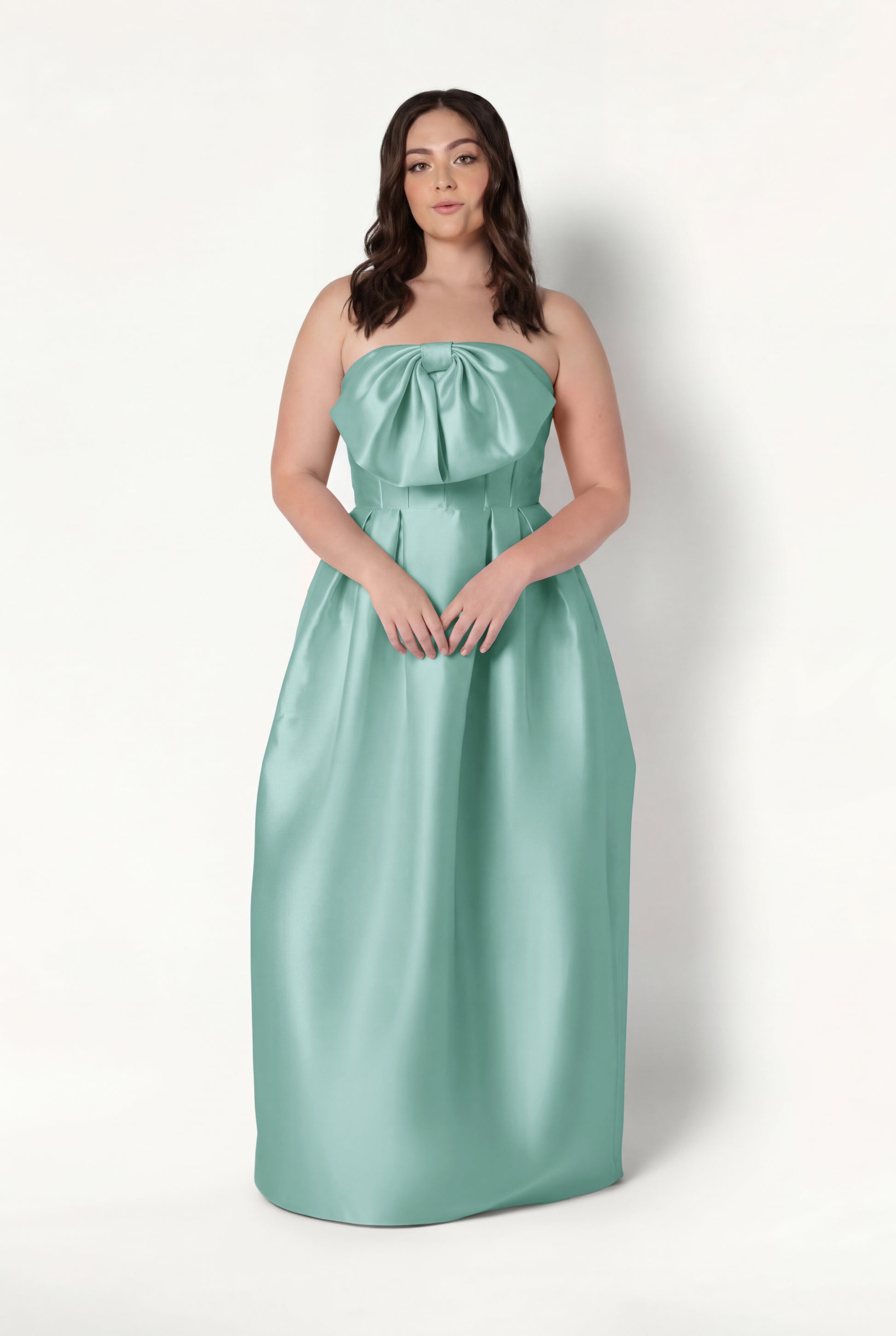 A woman with long, wavy brown hair and a neutral expression wears the TH&TH Ophelia Bridesmaid Dress in Seafoam Satin Twill—a strapless, floor-length gown featuring a corset bodice and large bow—against a plain white background.