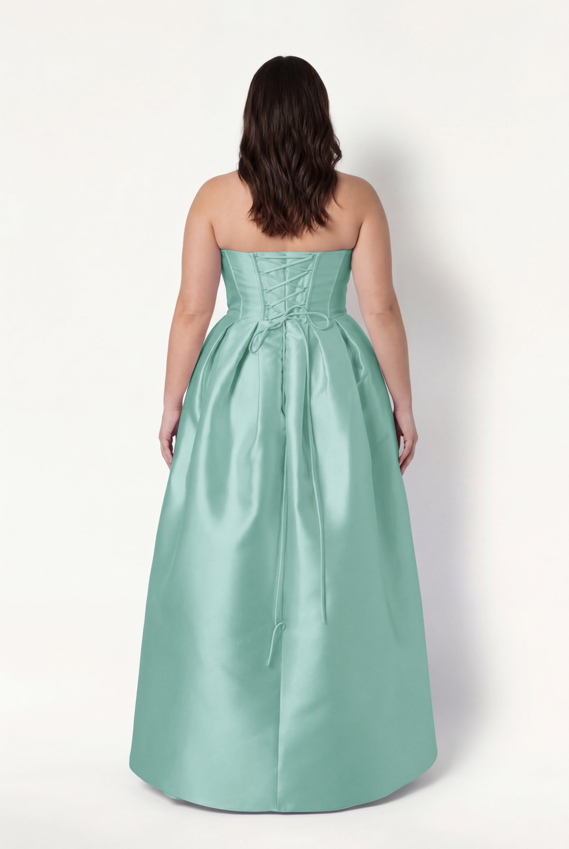 A woman with wavy brown hair faces away, wearing the TH&TH Ophelia Bridesmaid Dress in Seafoam Satin Twill—a strapless, floor-length gown featuring a lace-up corset bodice and full skirt.