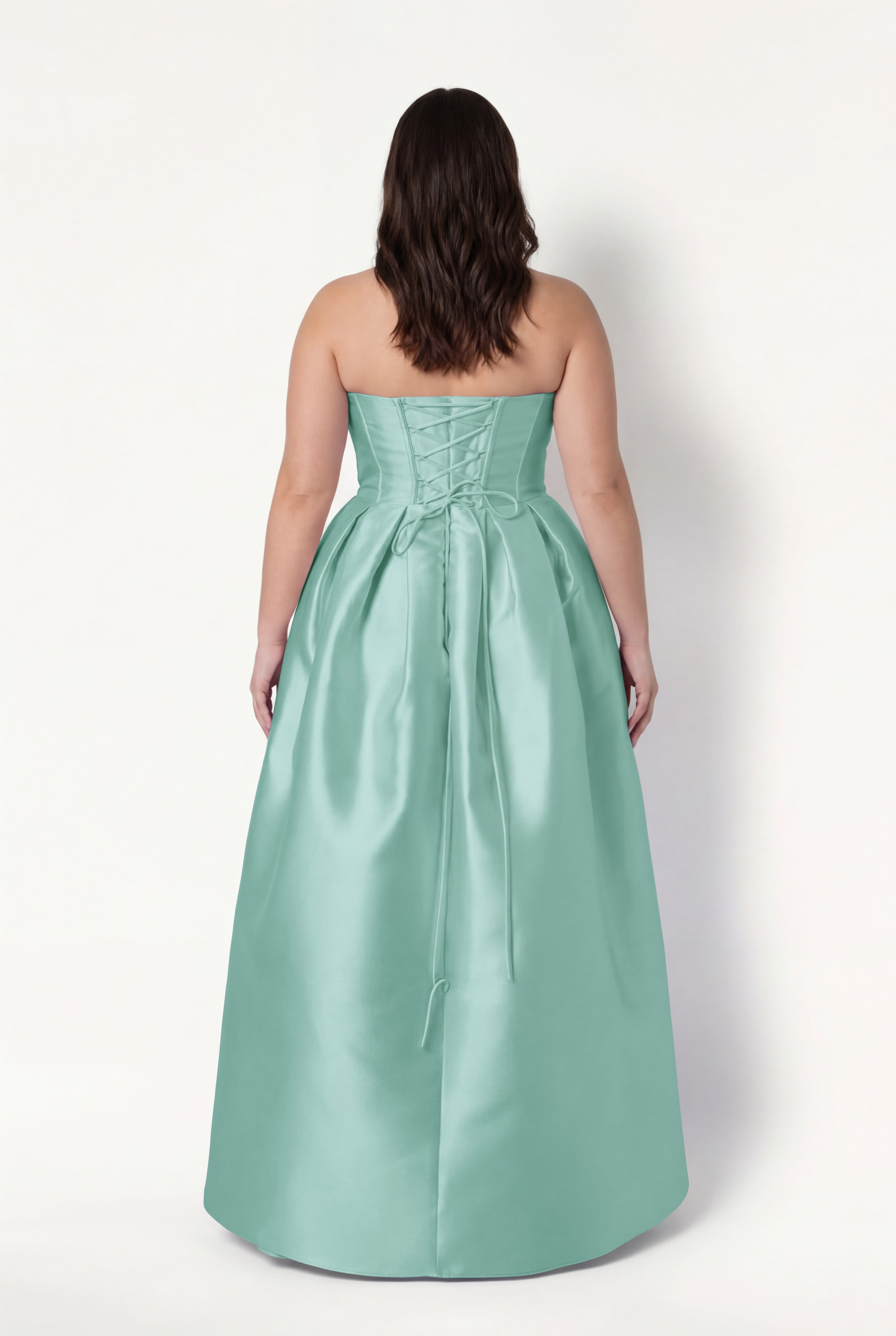 A woman with wavy brown hair faces away, wearing the TH&TH Ophelia Bridesmaid Dress in Seafoam Satin Twill—a strapless, floor-length gown featuring a lace-up corset bodice and full skirt.