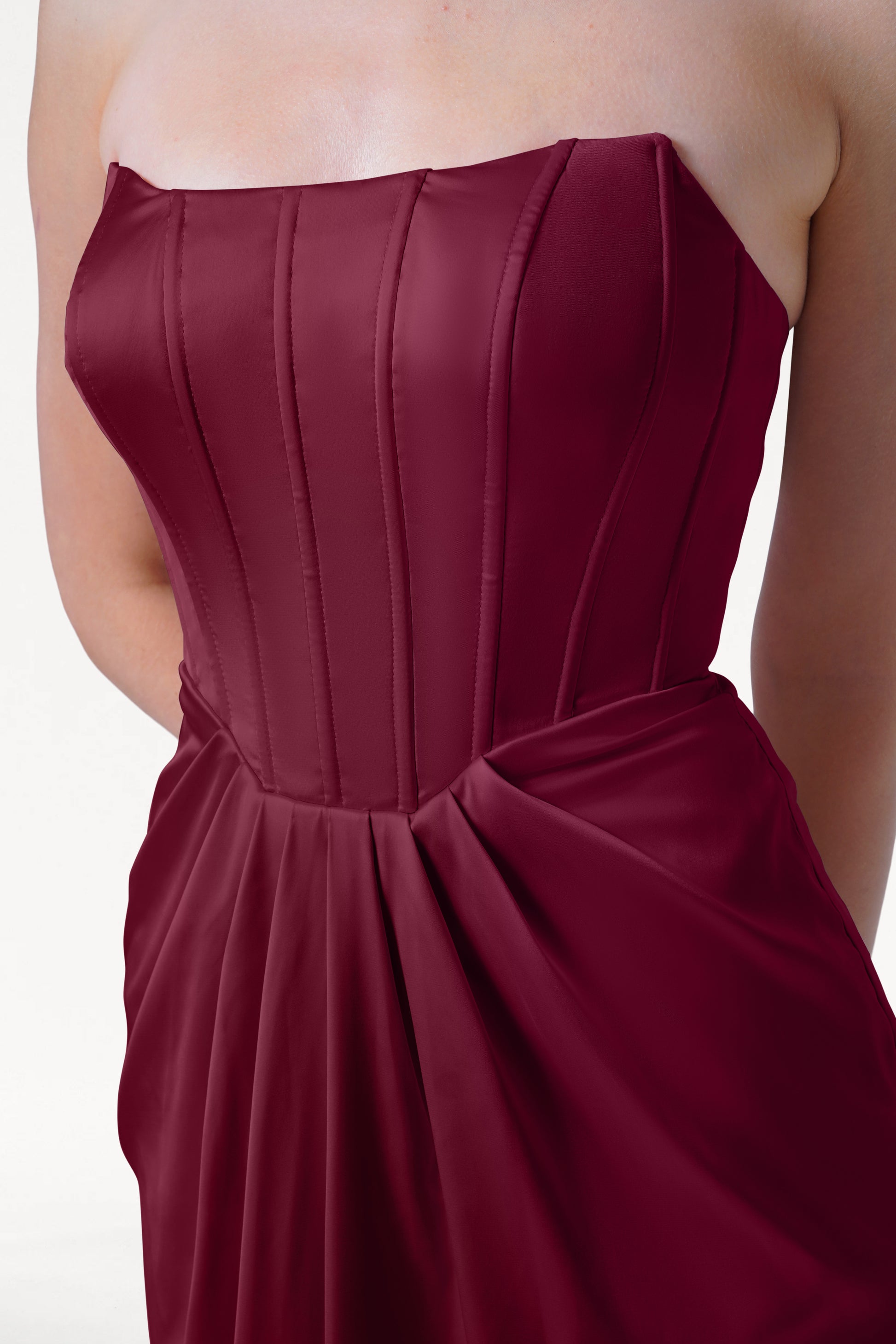 A woman stands against a plain white background, wearing the TH&TH Selena Bridesmaid Dress in Merlot Satin, a strapless gown with a corset bodice, vertical seams, and pleated waist detail.
