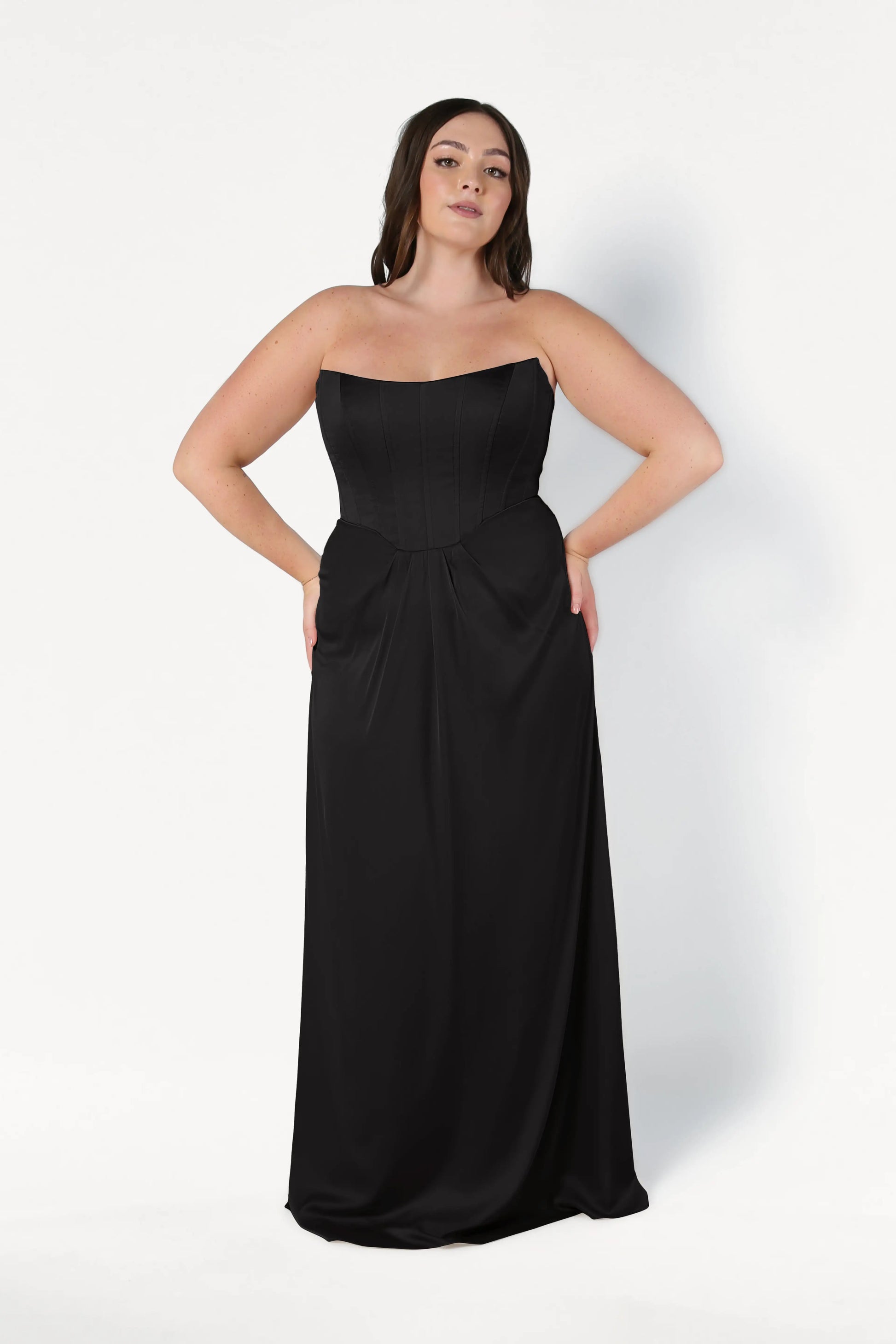 A woman with long dark hair wears the TH&TH Selena Bridesmaid Dress in Black Satin—a strapless, floor-length gown with a corset bodice and relaxed skirt—standing against a plain white background.