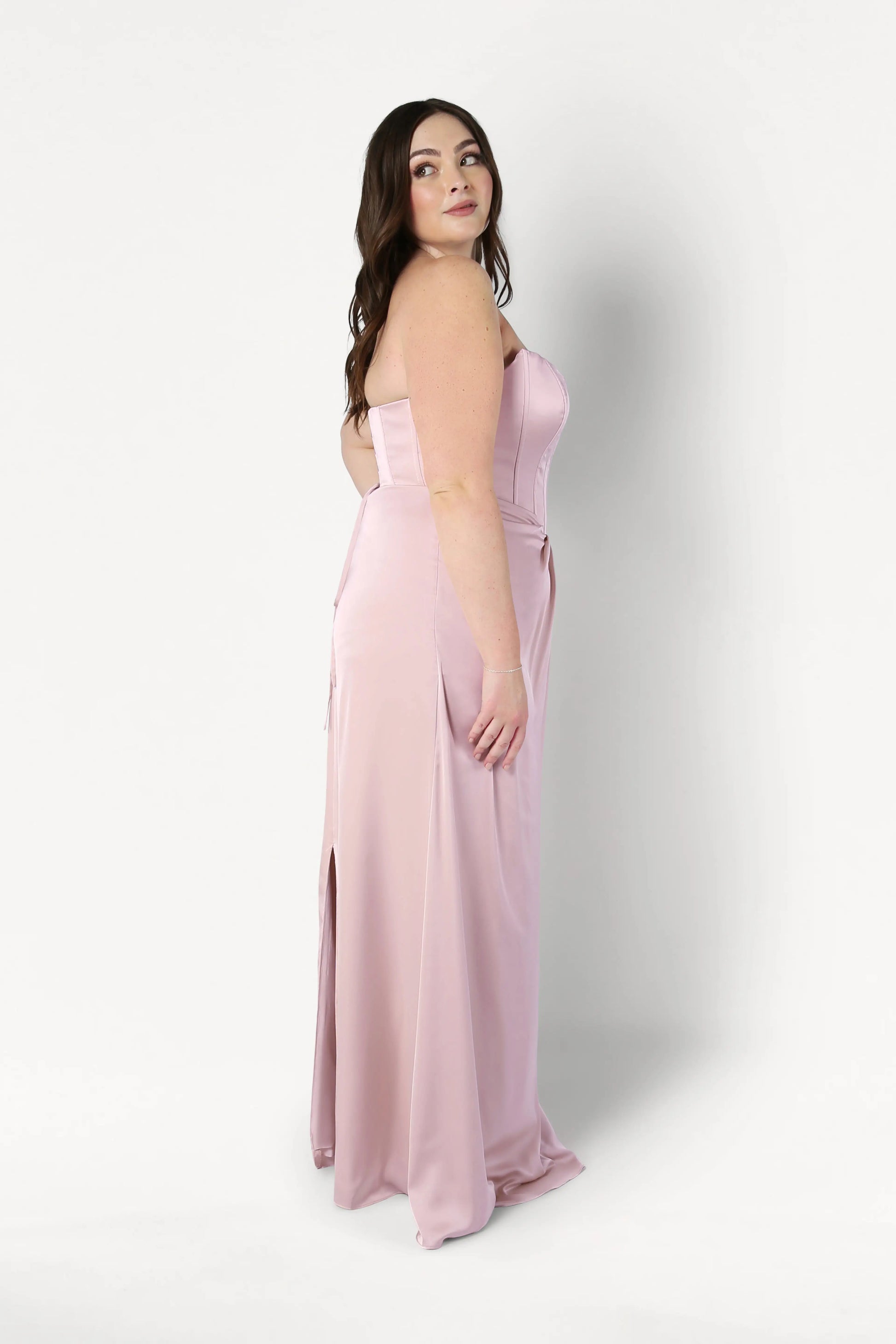 A woman with long brown hair models the size-inclusive Selena Bridesmaid Dress in Blush Pink Satin by TH&TH. The dress features a corset bodice and high slit. She stands in profile against a plain white background, looking over her shoulder.