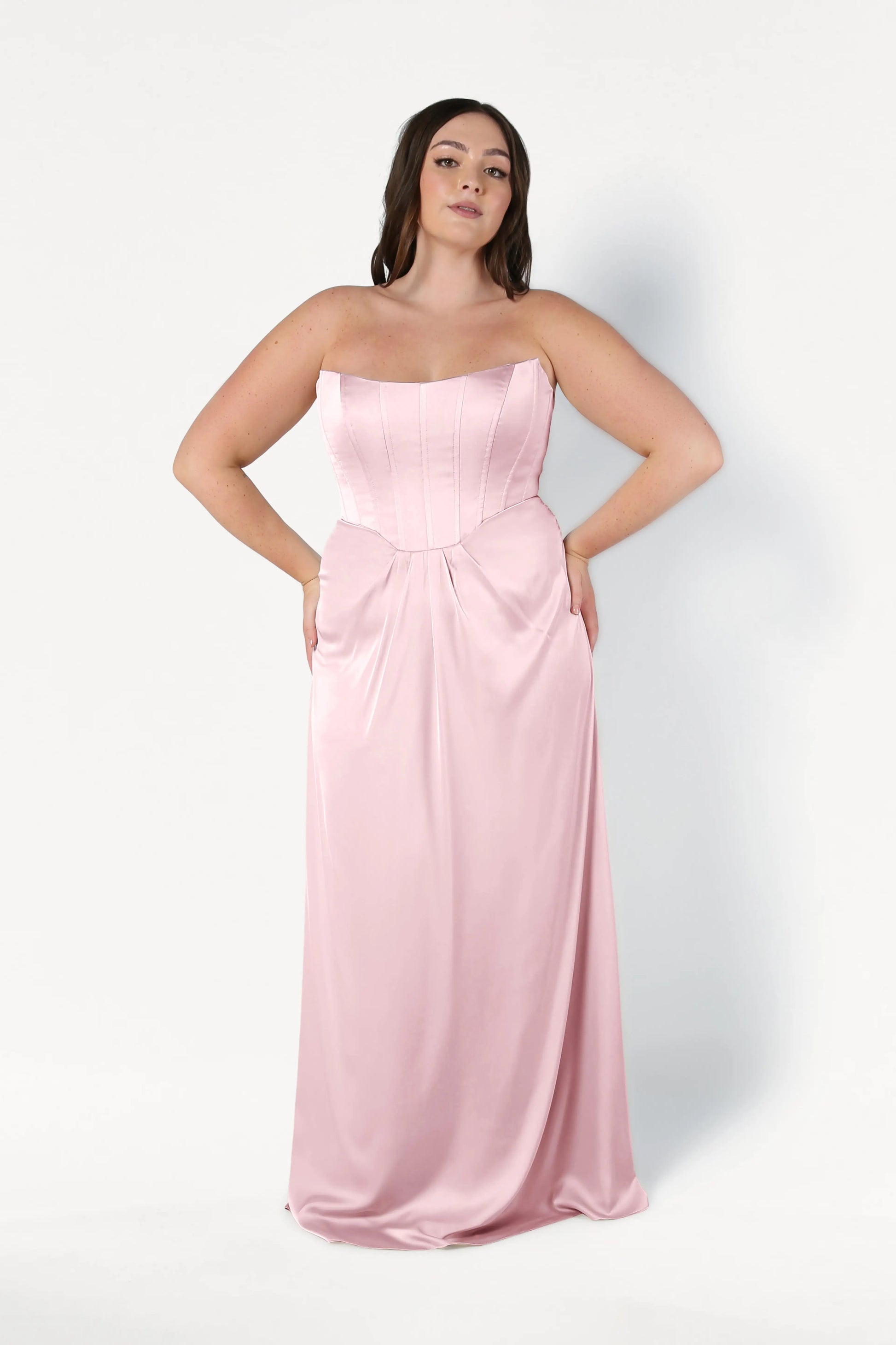 A woman with long dark hair poses against a white background wearing the TH&TH Selena Bridesmaid Dress in blush pink satin, featuring a strapless fitted corset bodice and flowing skirt, with her hands on her hips.