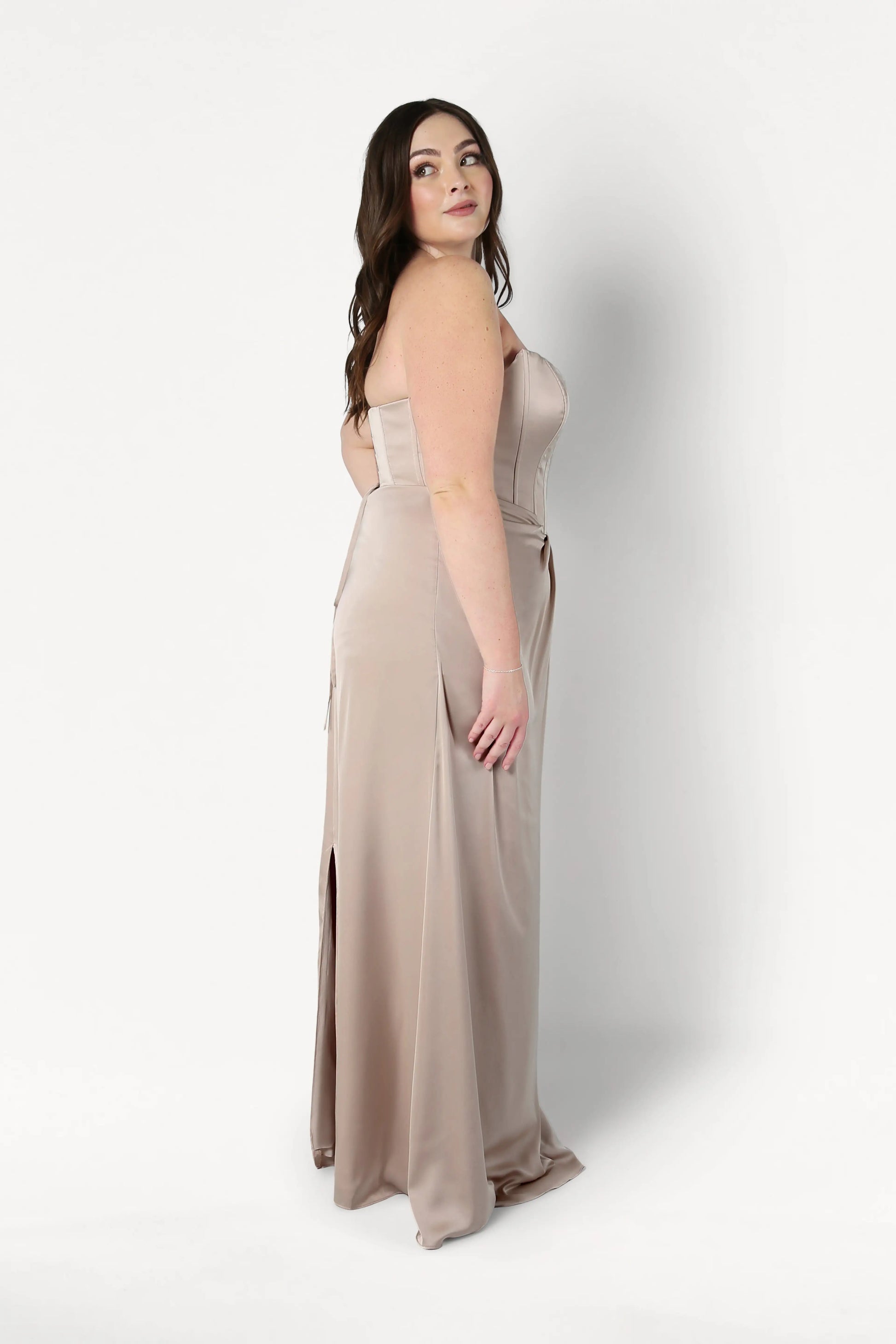 A woman with long brown hair models the Selena Bridesmaid Dress in Champagne Sky Satin by TH&TH, a size-inclusive strapless gown, standing sideways and glancing over her shoulder against a plain white backdrop.