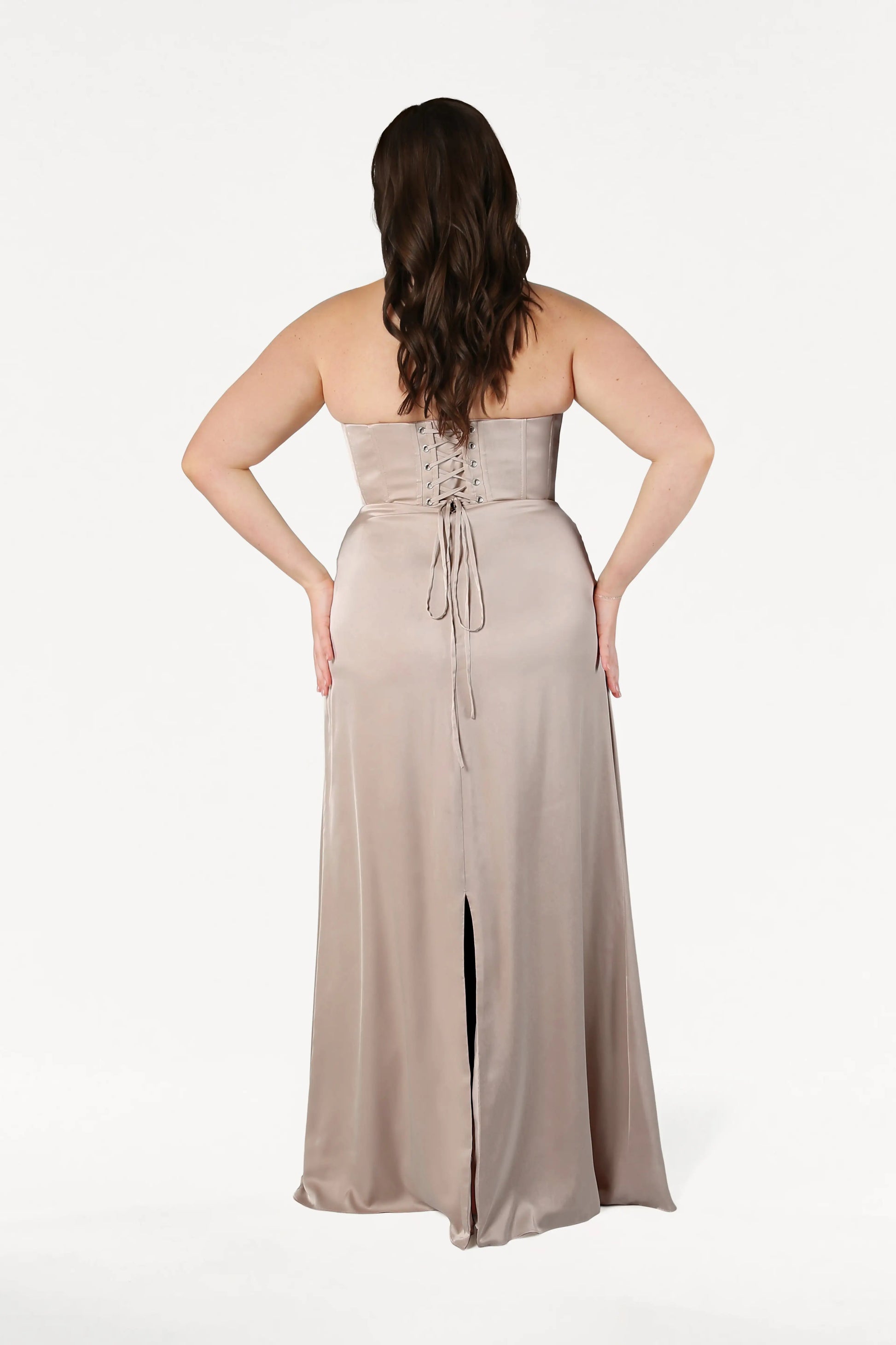 A woman faces away in the Selena Bridesmaid Dress by TH&TH, crafted from Champagne Sky Satin with a corset bodice, lace-up back, and hem slit. Her hands rest on her hips; wavy brown hair falls to her upper back. Background is white.