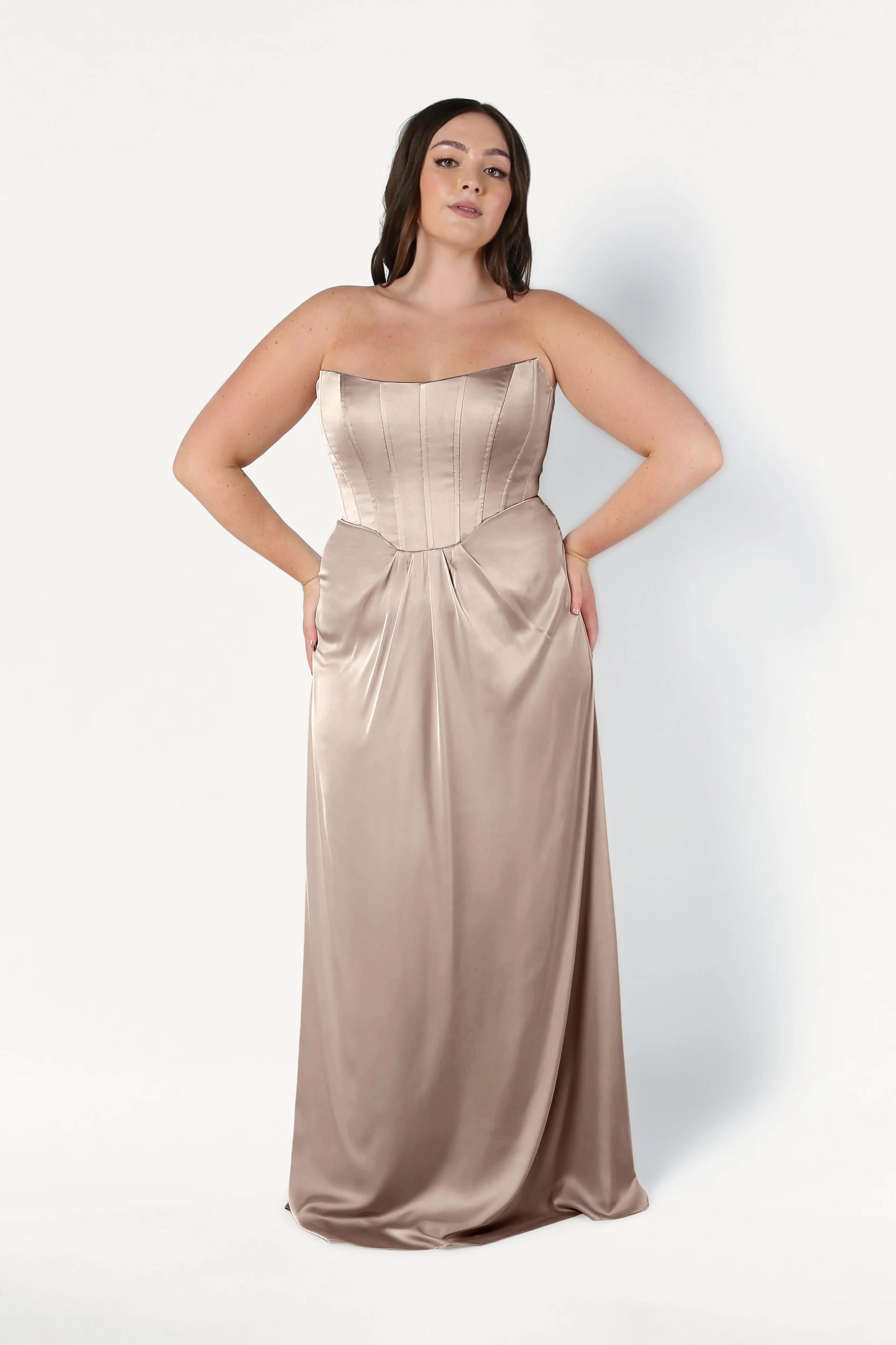 A woman poses against a white background in the TH&TH Selena Bridesmaid Dress in Champagne Sky Satin, featuring a strapless, floor-length silhouette with a fitted corset bodice and subtle sheen, her hair loosely styled.