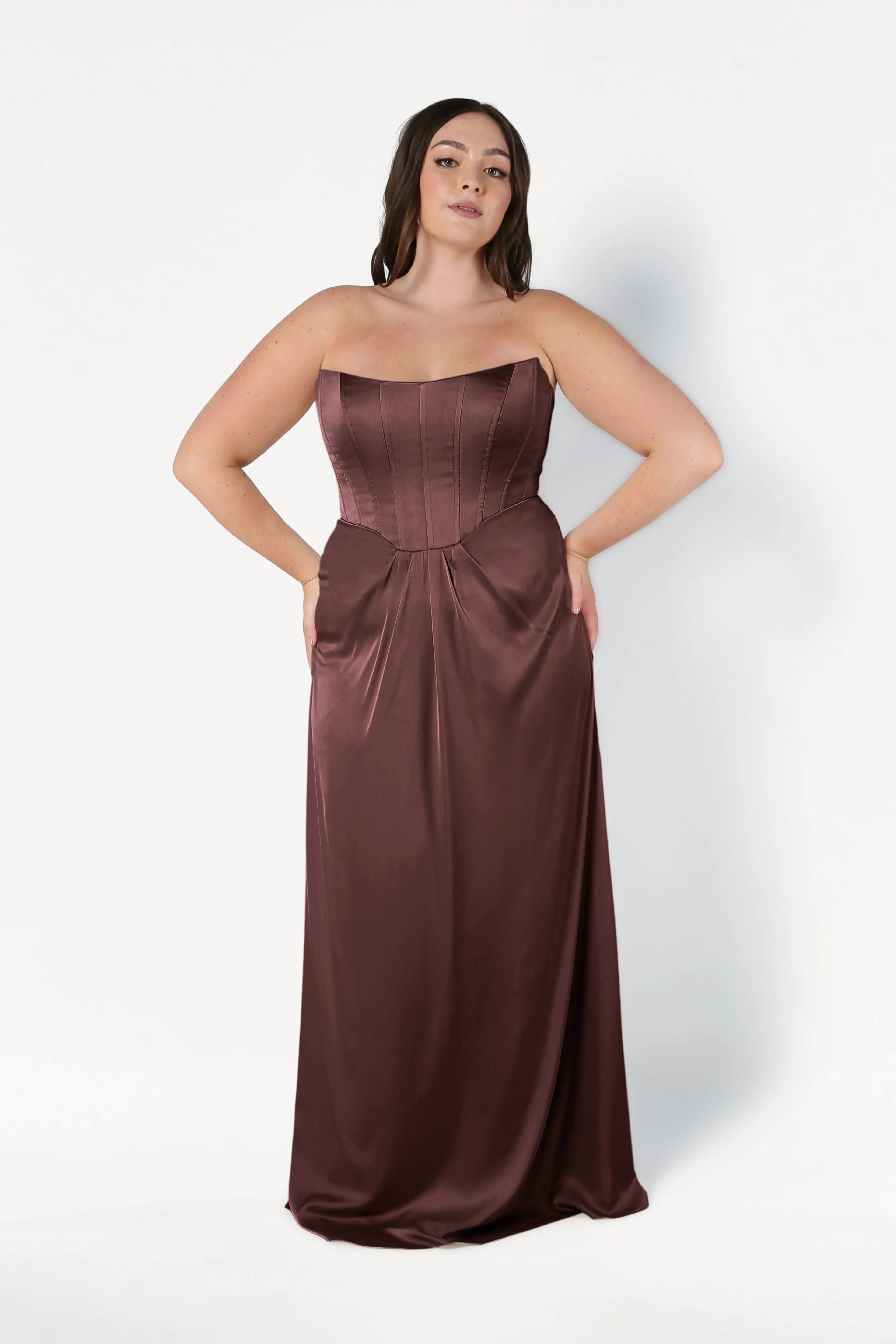 A woman stands against a white background, hands in the pockets of the TH&TH Selena Bridesmaid Dress in Chocolate Brown Satin, featuring a strapless floor-length design with a corset bodice and subtle draped pleats.