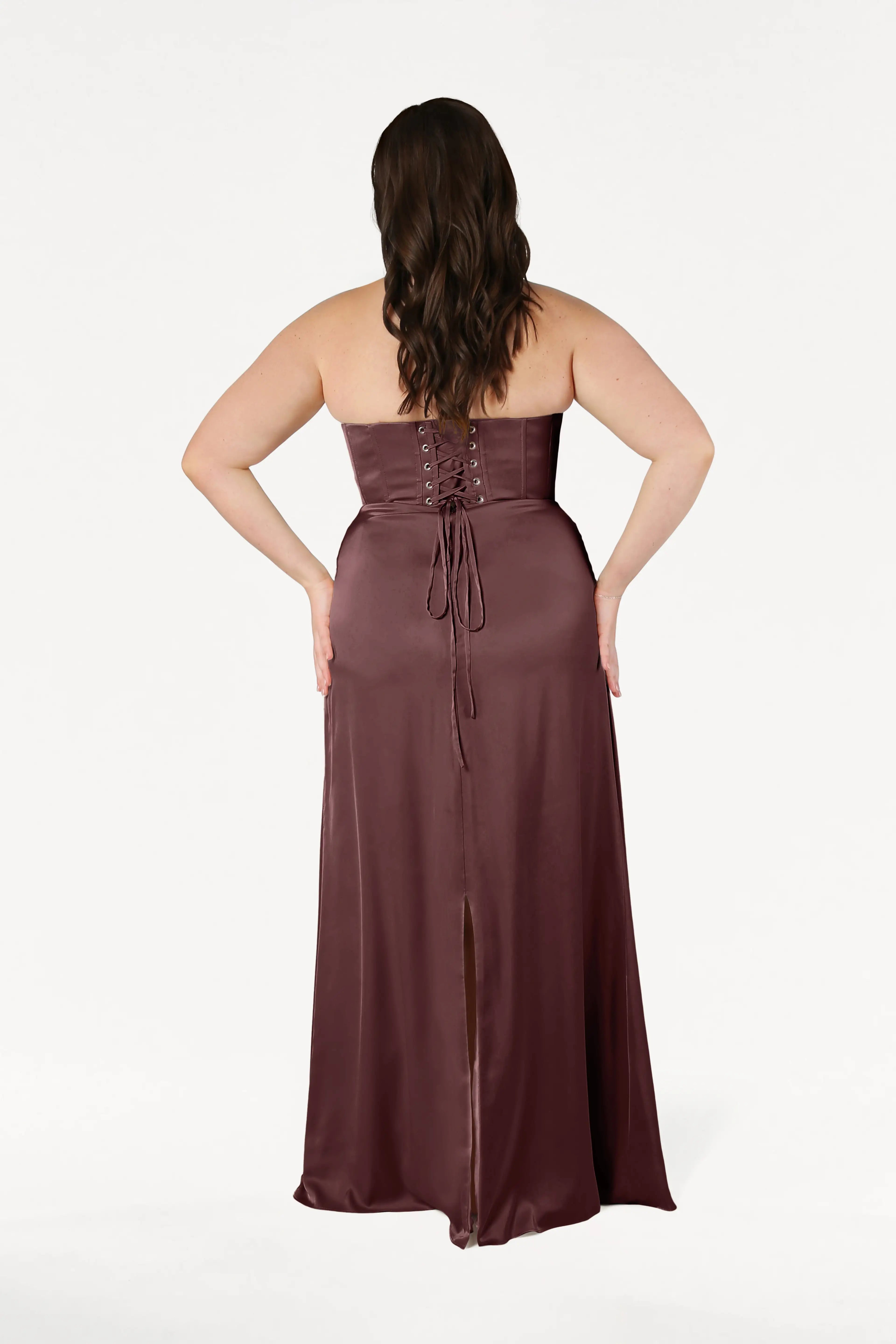 A woman with wavy brown hair faces away from the camera, wearing the TH&TH Selena Bridesmaid Dress in Chocolate Brown Satin—strapless, floor-length, with draped pleats, corset bodice, lace-up back, and a center slit against a white background.