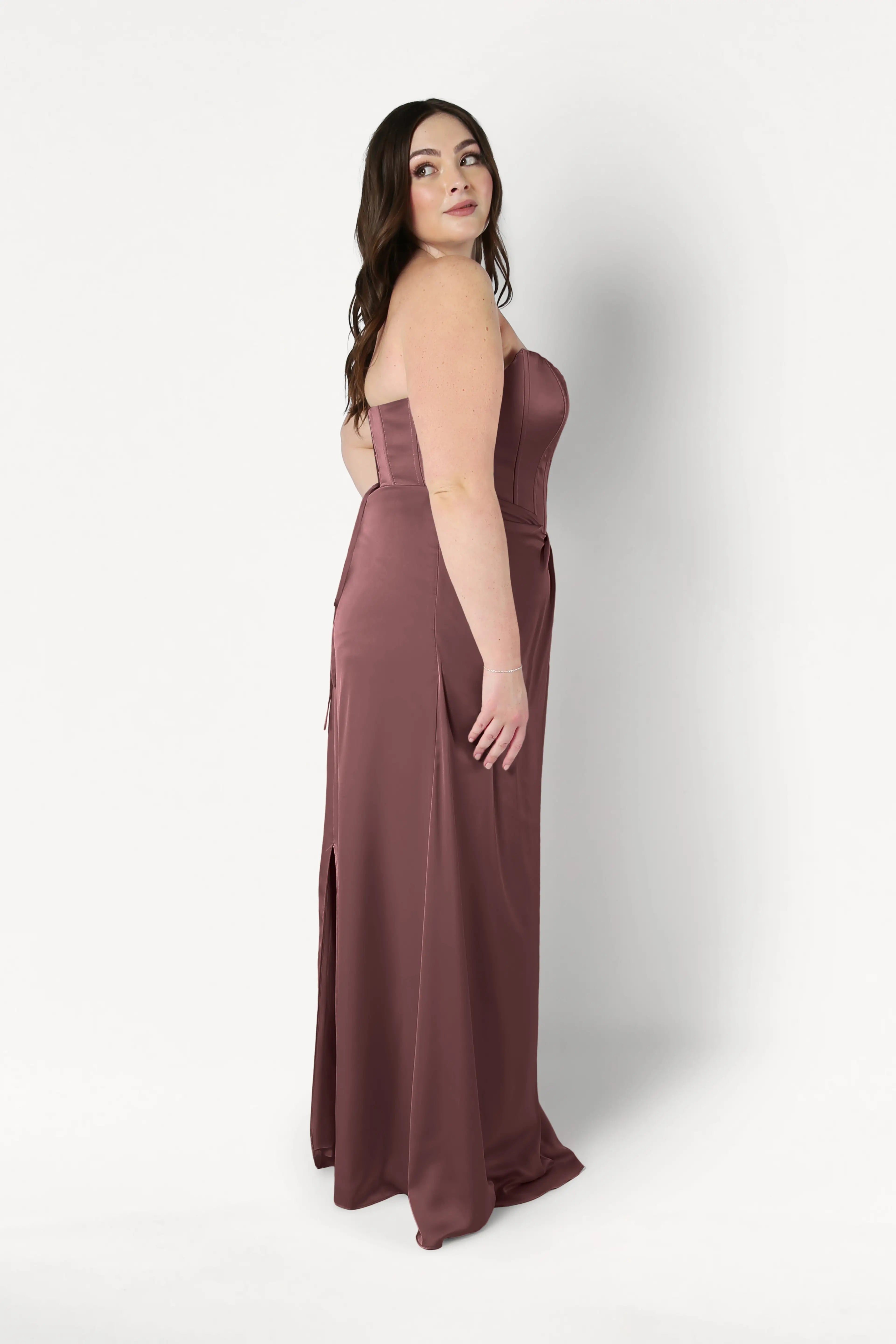 A woman with long brown hair wears the TH&TH Selena Bridesmaid Dress in chocolate brown satin. The strapless, floor-length gown features draped pleats and a side slit as she poses sideways against a plain white background.
