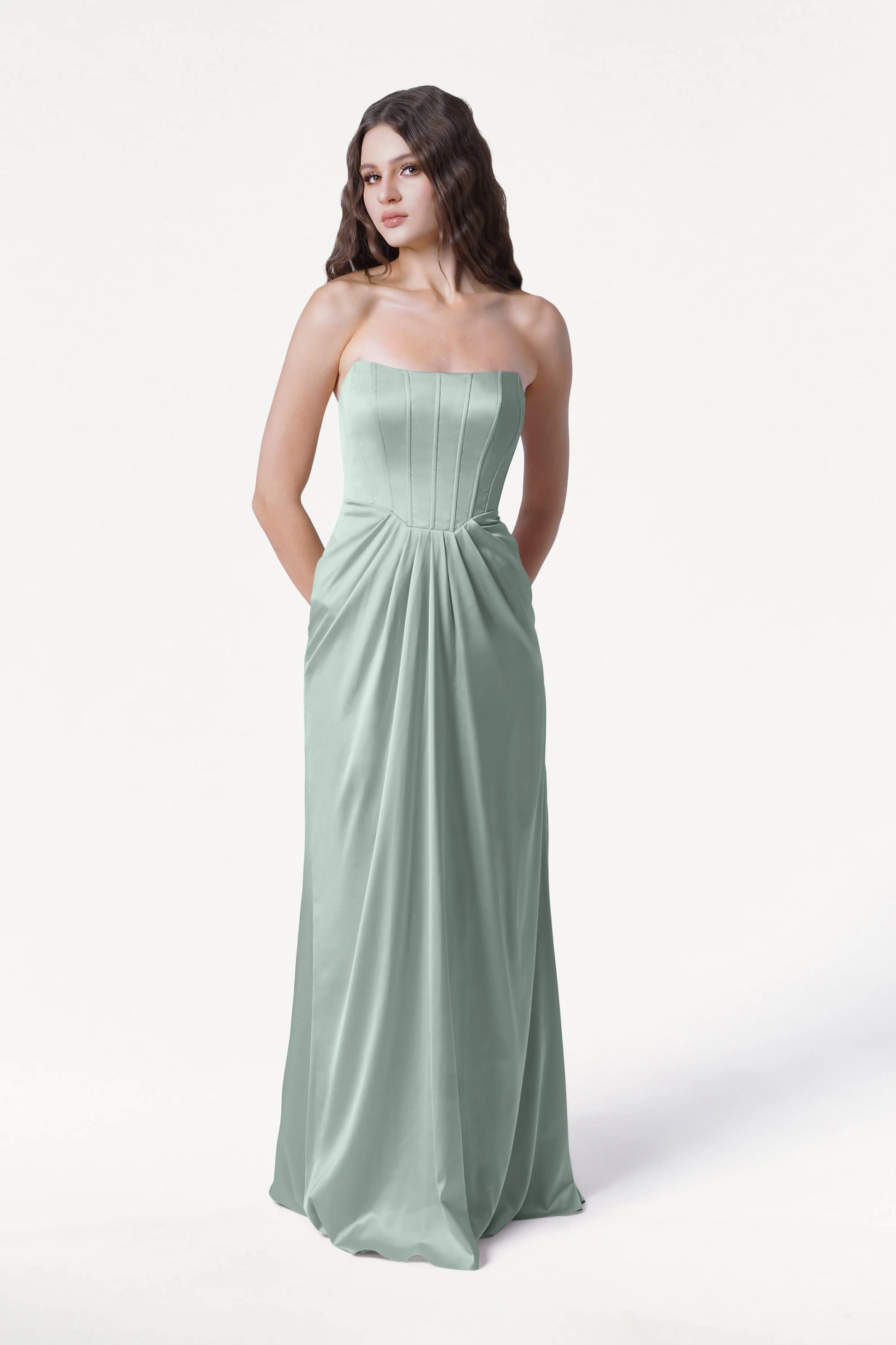 A woman poses against a white background wearing the TH&TH Selena Bridesmaid Dress in Dusty Eucalyptus Satin, featuring a strapless, floor-length design with pleated detailing, corset bodice, and pockets.