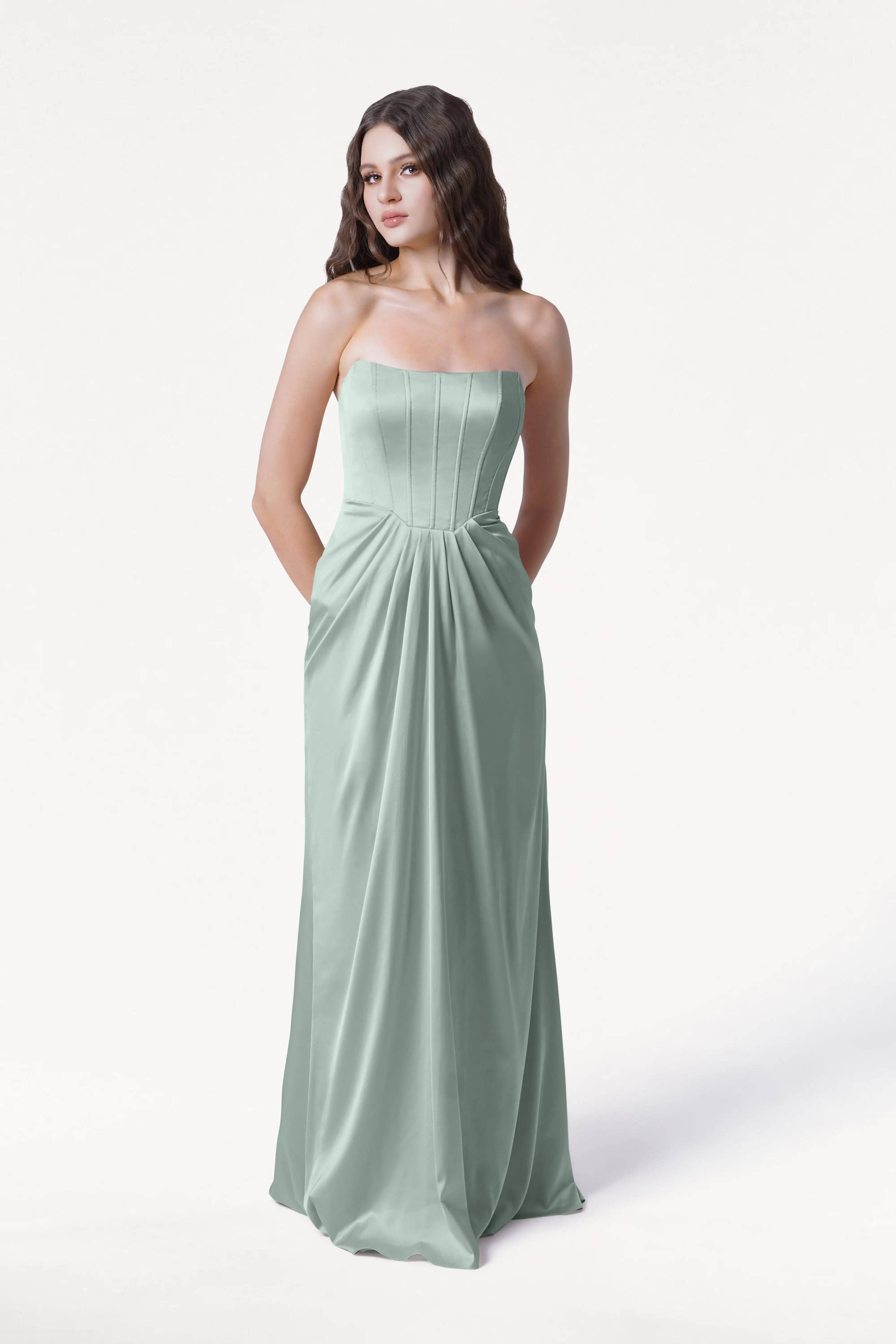 A woman poses against a white background wearing the TH&TH Selena Bridesmaid Dress in Dusty Eucalyptus Satin, featuring a strapless, floor-length design with pleated detailing, corset bodice, and pockets.