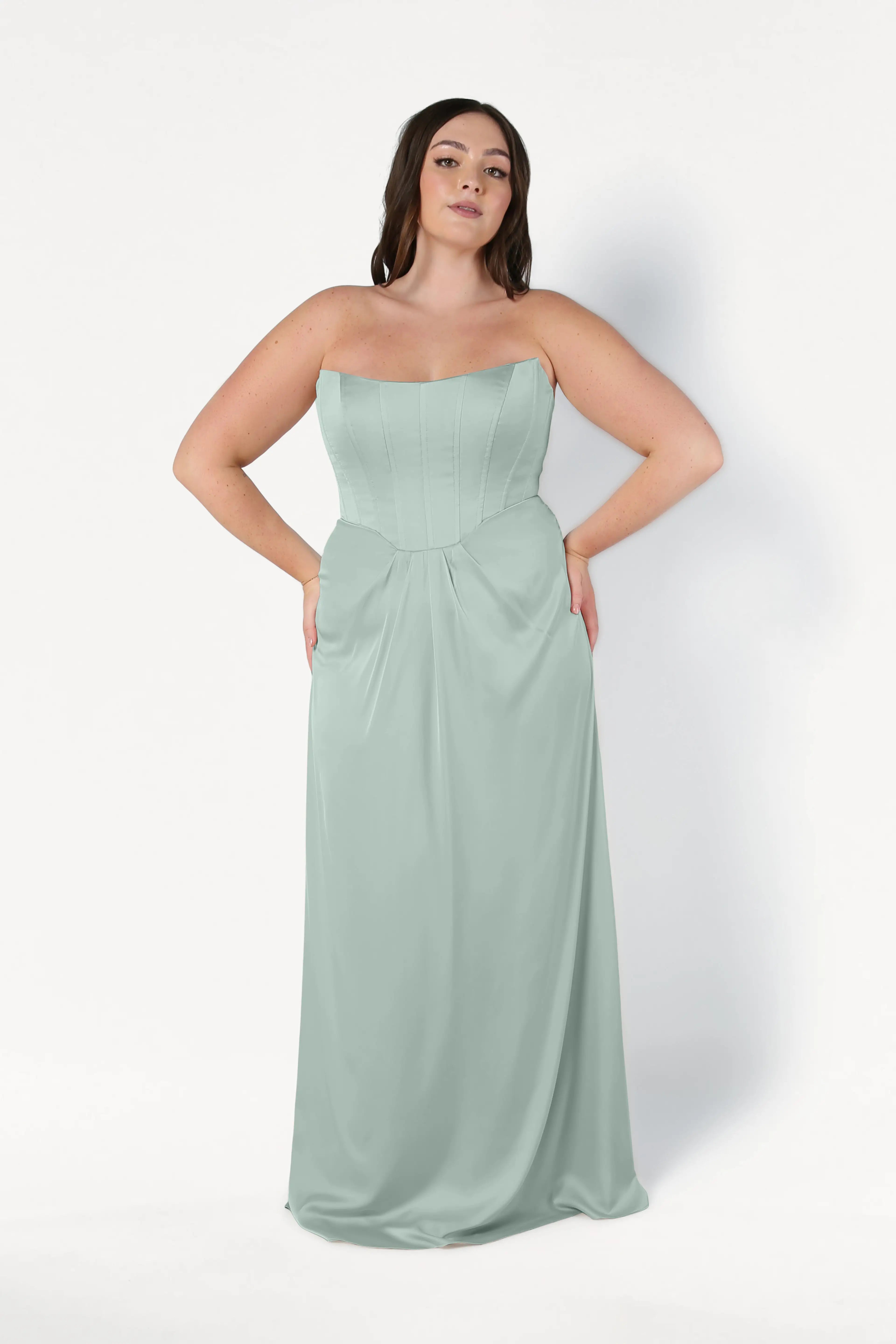 A woman with long, dark hair stands against a white background in the TH&TH Selena Bridesmaid Dress in Dusty Eucalyptus Satin, a size-inclusive strapless gown with a fitted corset bodice and flowing floor-length skirt.