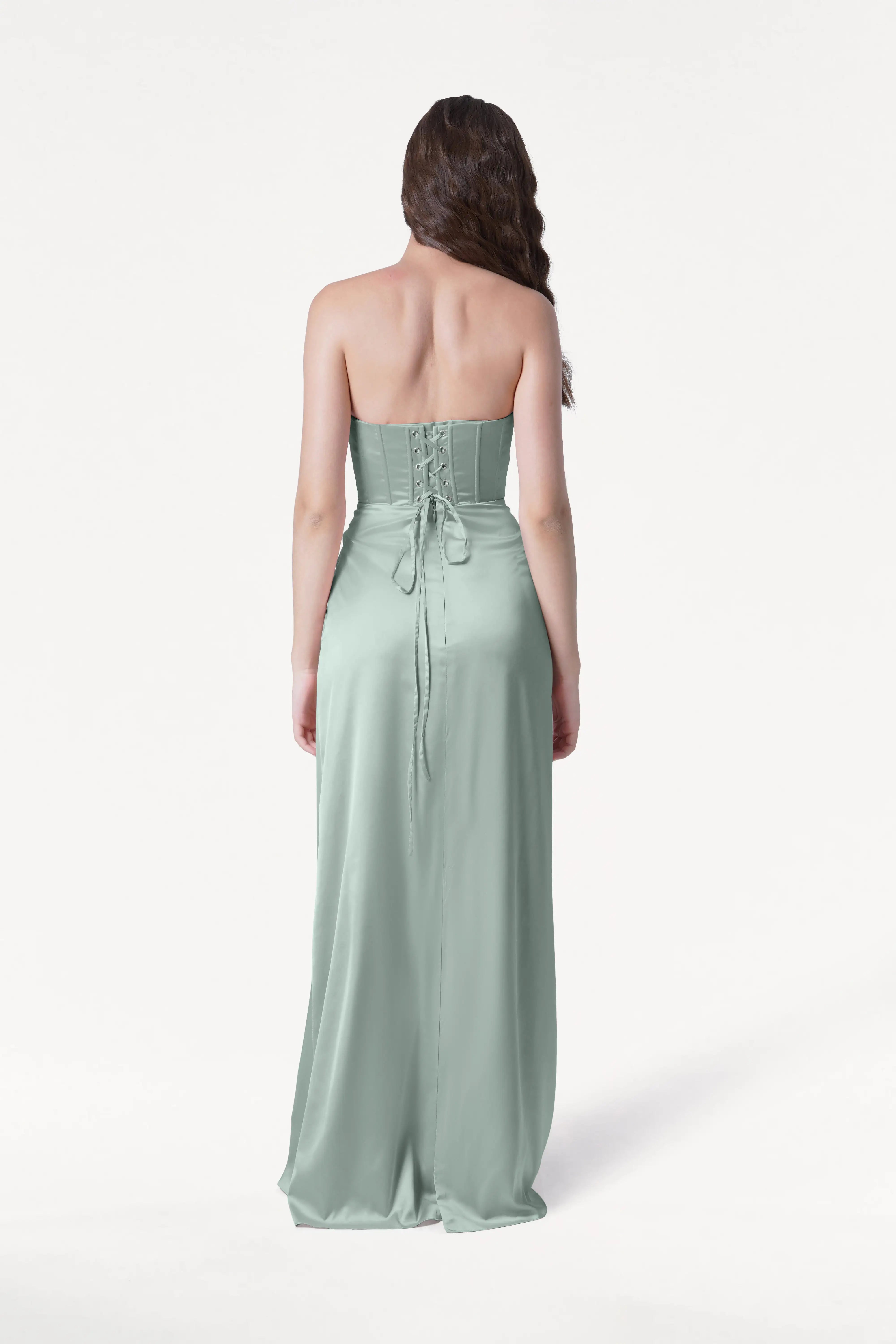 A woman faces away, wearing the TH&TH Selena Bridesmaid Dress in Dusty Eucalyptus Satin. This size-inclusive gown features a corset bodice, lace-up back, and long flowing skirt. Her wavy brown hair drapes over one shoulder against a white background.