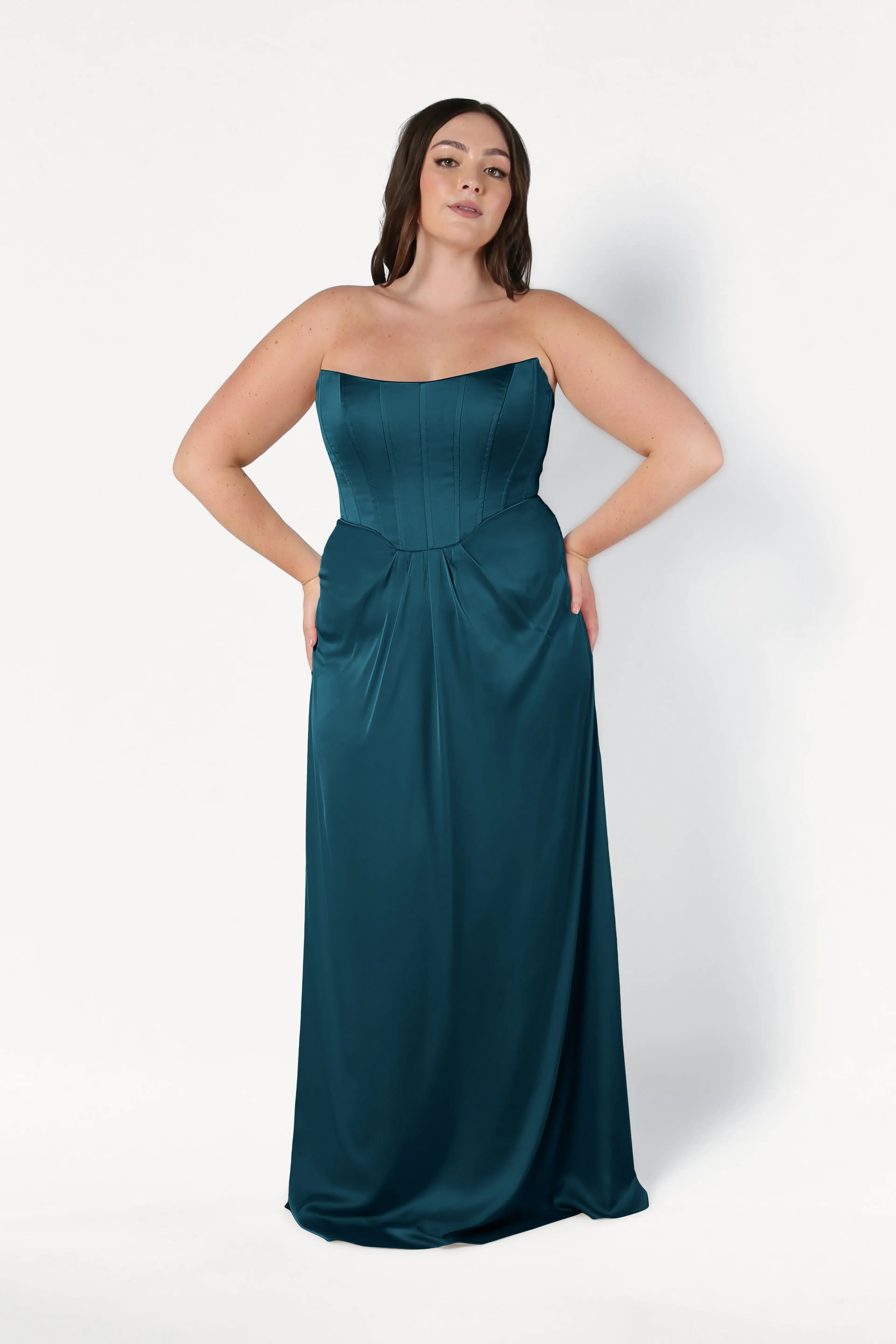 A woman with long dark hair models the TH&TH Selena Bridesmaid Dress in Emerald Dawn Satin, featuring a strapless fitted corset bodice and flowing skirt, posing with hands on her hips against a plain white background.