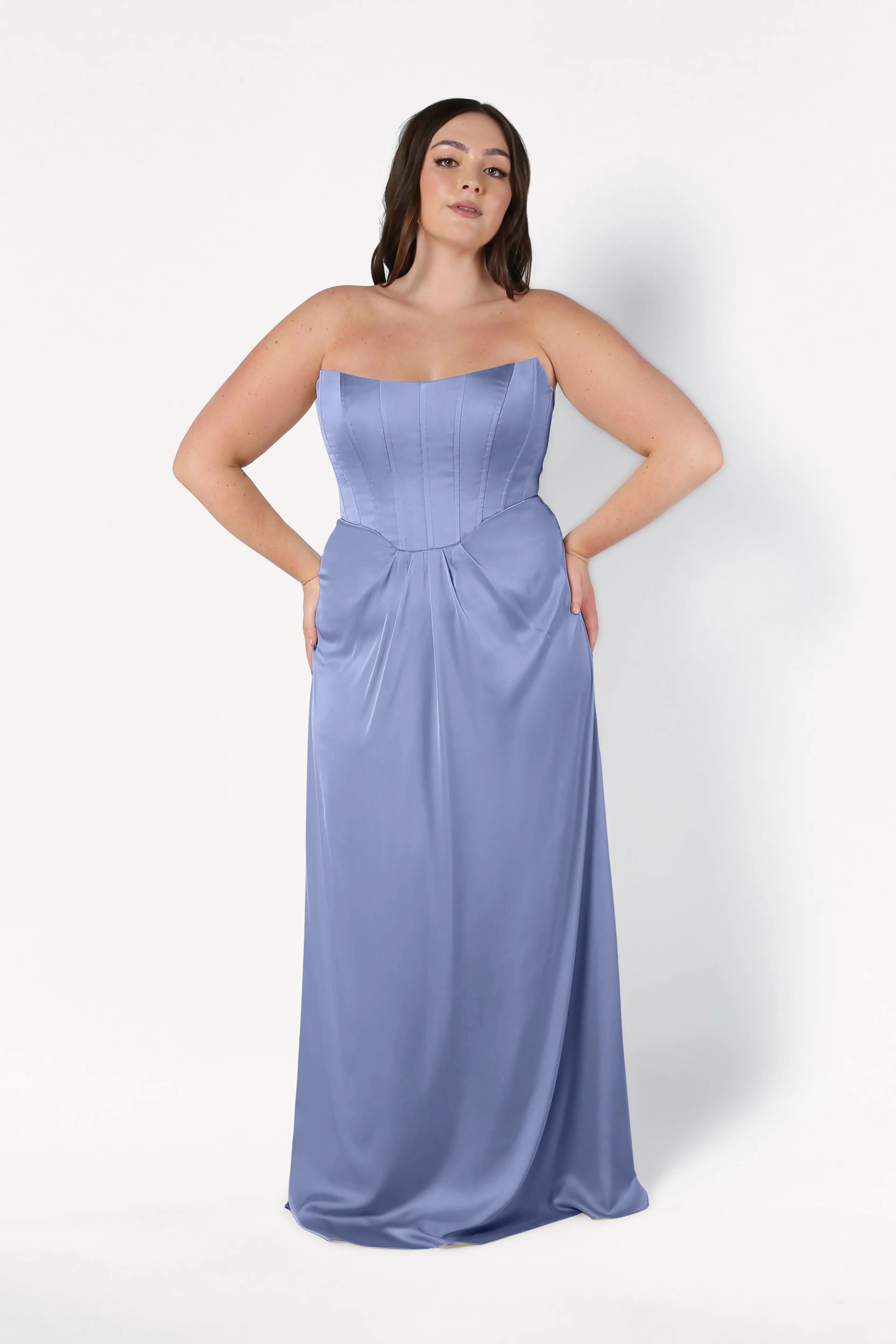 A woman with long dark hair stands against a white background wearing the TH&TH Selena Bridesmaid Dress in French Blue Satin, a strapless, floor-length gown with elegant draped pleats and her hands on her hips.