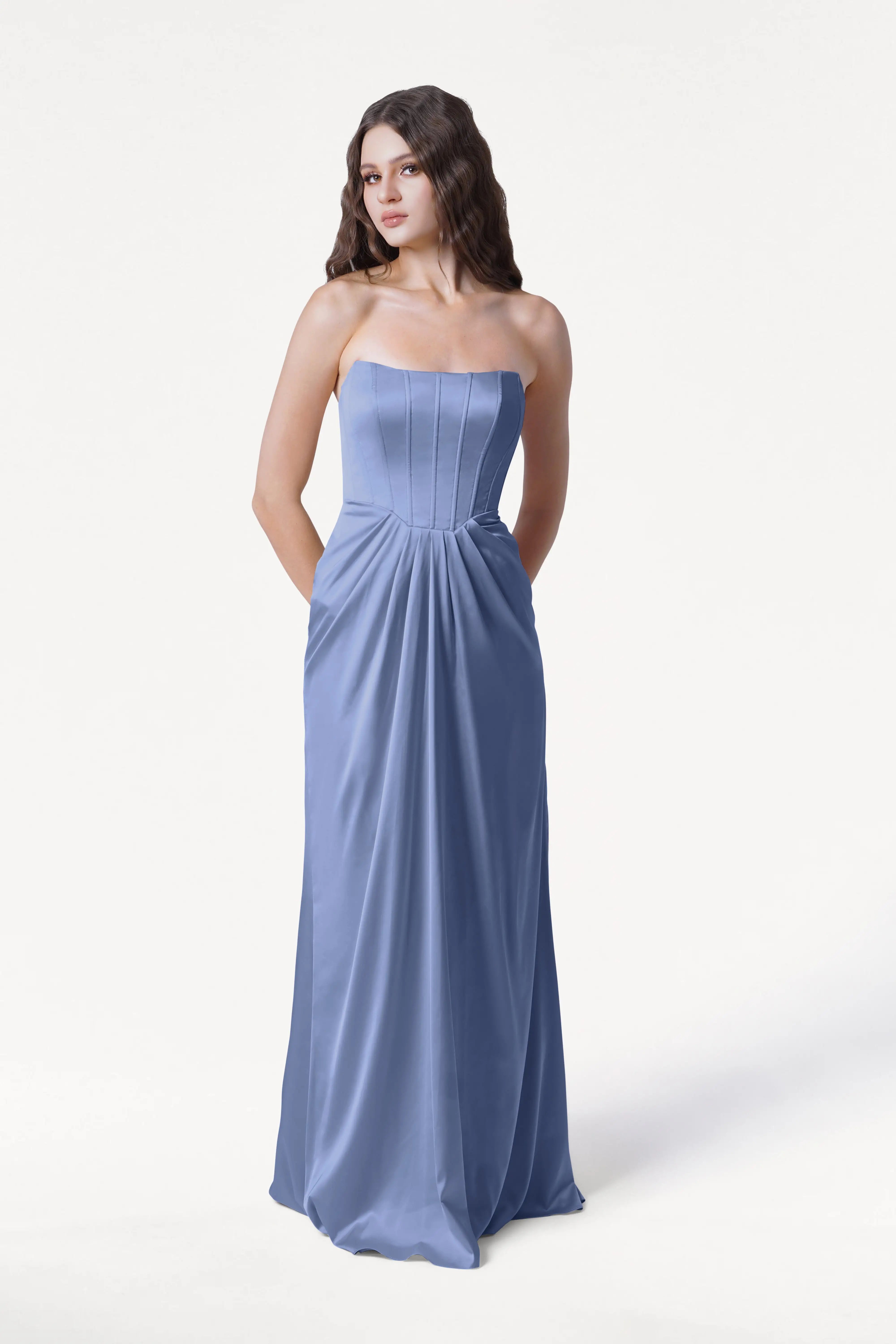 A woman stands against a plain white background wearing the TH&TH Selena Bridesmaid Dress in French Blue Satin, a strapless, floor-length gown with draped pleats and pockets.