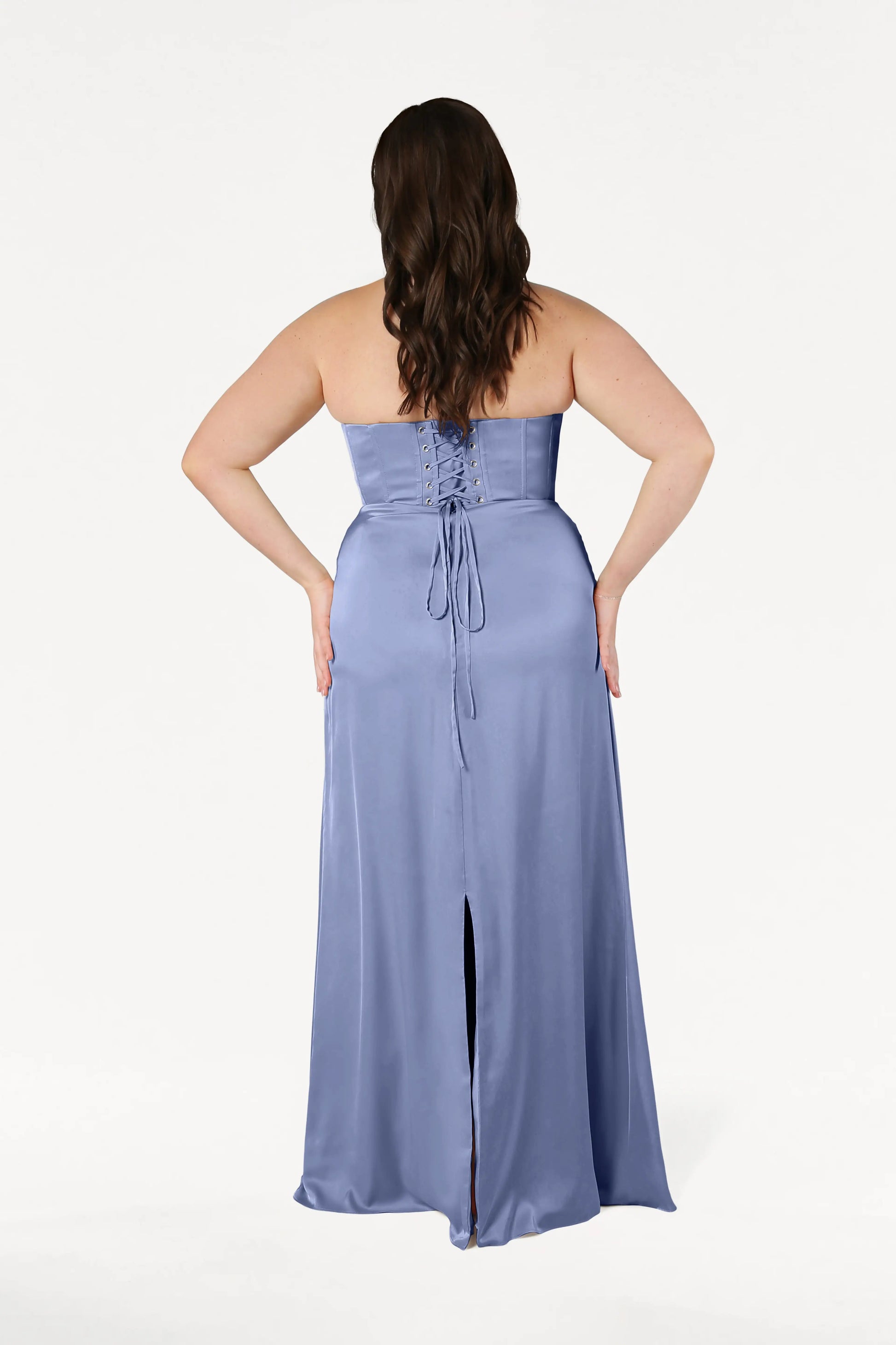 A woman with long, wavy brown hair is shown from the back wearing the Selena Bridesmaid Dress in French Blue Satin by TH&TH. The strapless design features draped pleats, a lace-up corset bodice, back slit, and floor-length silhouette on a plain white background.