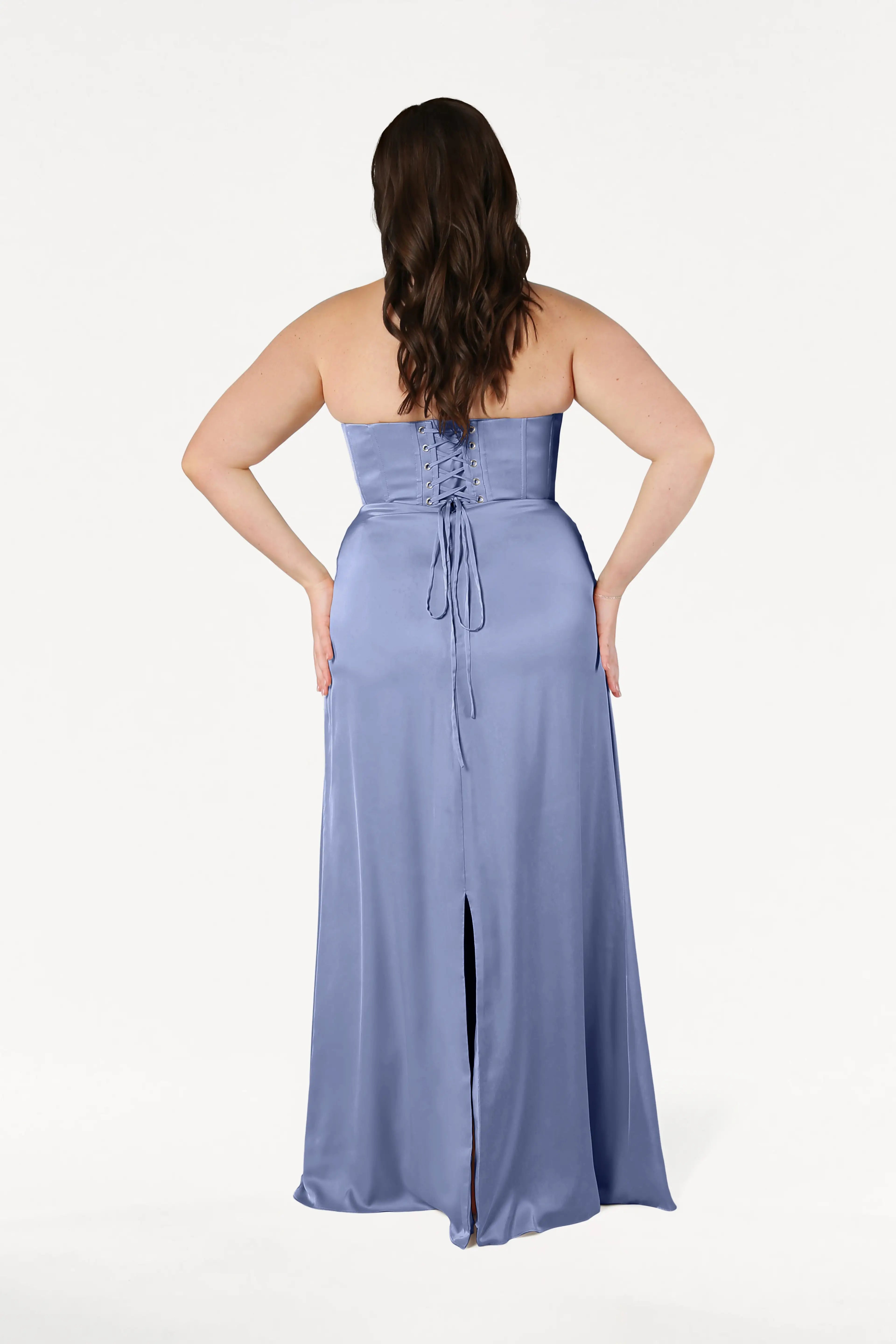A woman with long, wavy brown hair is shown from the back wearing the Selena Bridesmaid Dress in French Blue Satin by TH&TH. The strapless design features draped pleats, a lace-up corset bodice, back slit, and floor-length silhouette on a plain white background.
