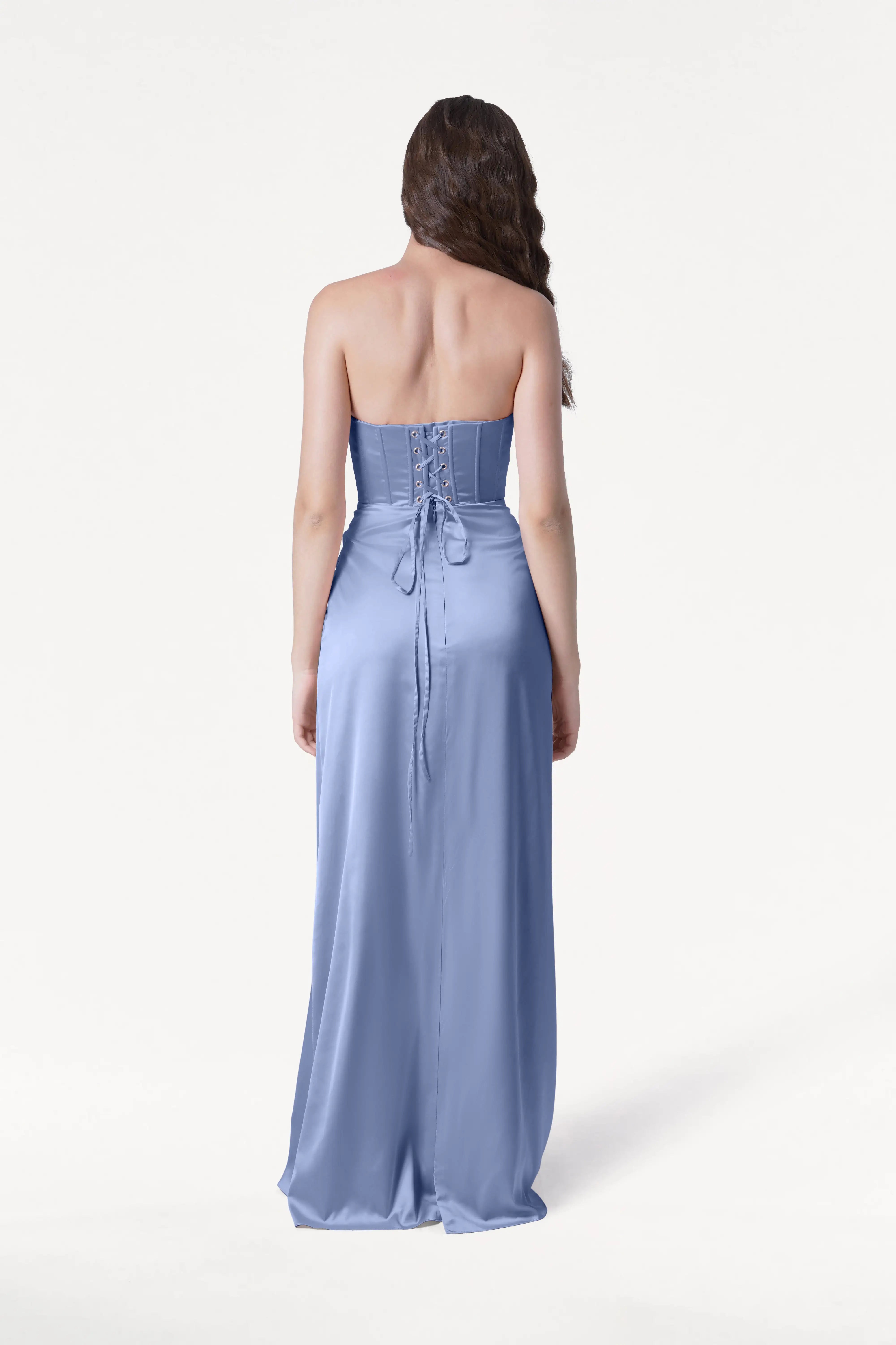 A woman with long brown hair faces away in the TH&TH Selena Bridesmaid Dress in French Blue Satin, a strapless, floor-length gown with draped pleats and a corset-style lace-up back, set against a plain white background.