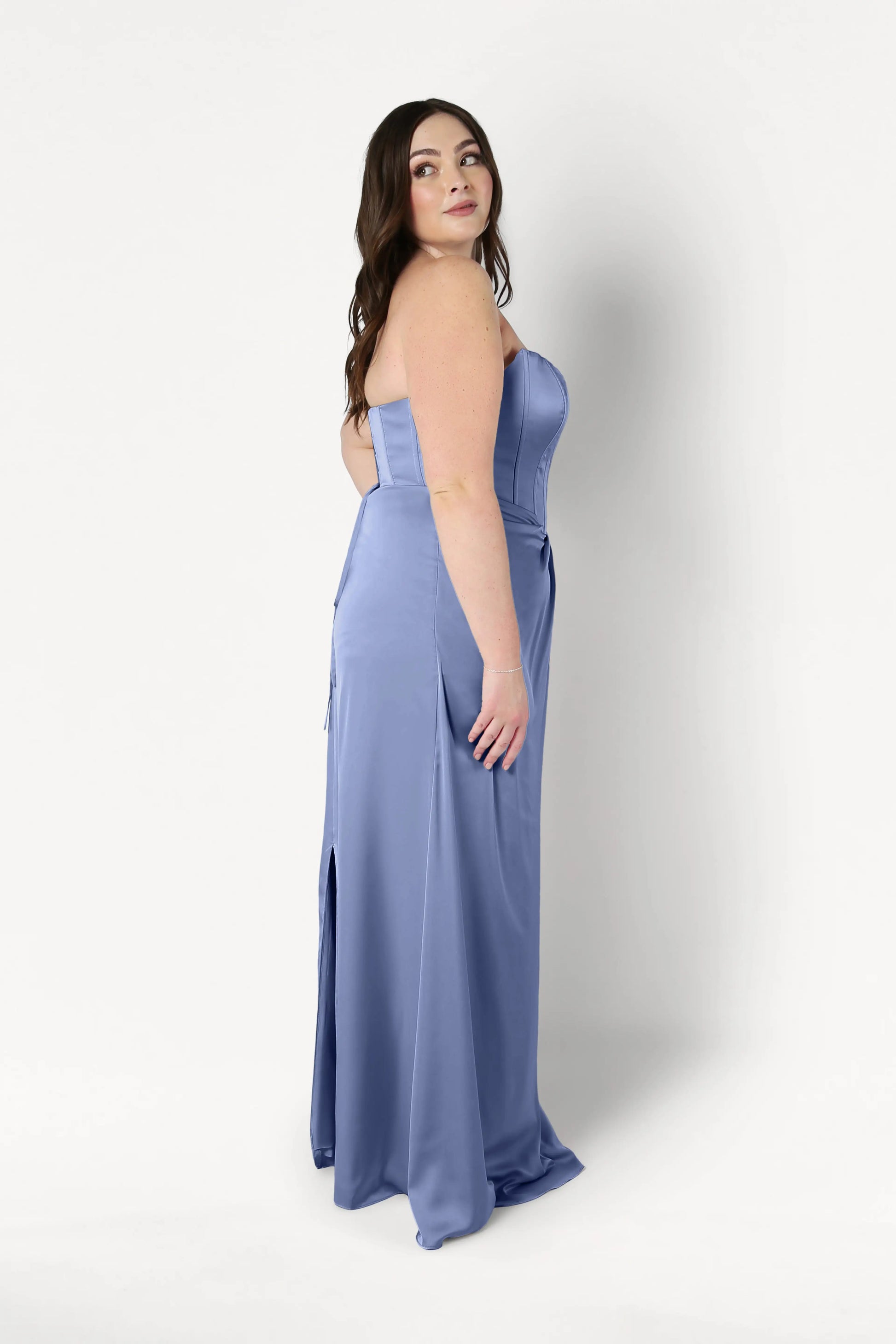 A woman with long brown hair wears the Selena Bridesmaid Dress in French Blue Satin by TH&TH. The strapless, floor-length gown features a corset bodice, draped pleats, and a side slit as she poses in profile against a plain white background.