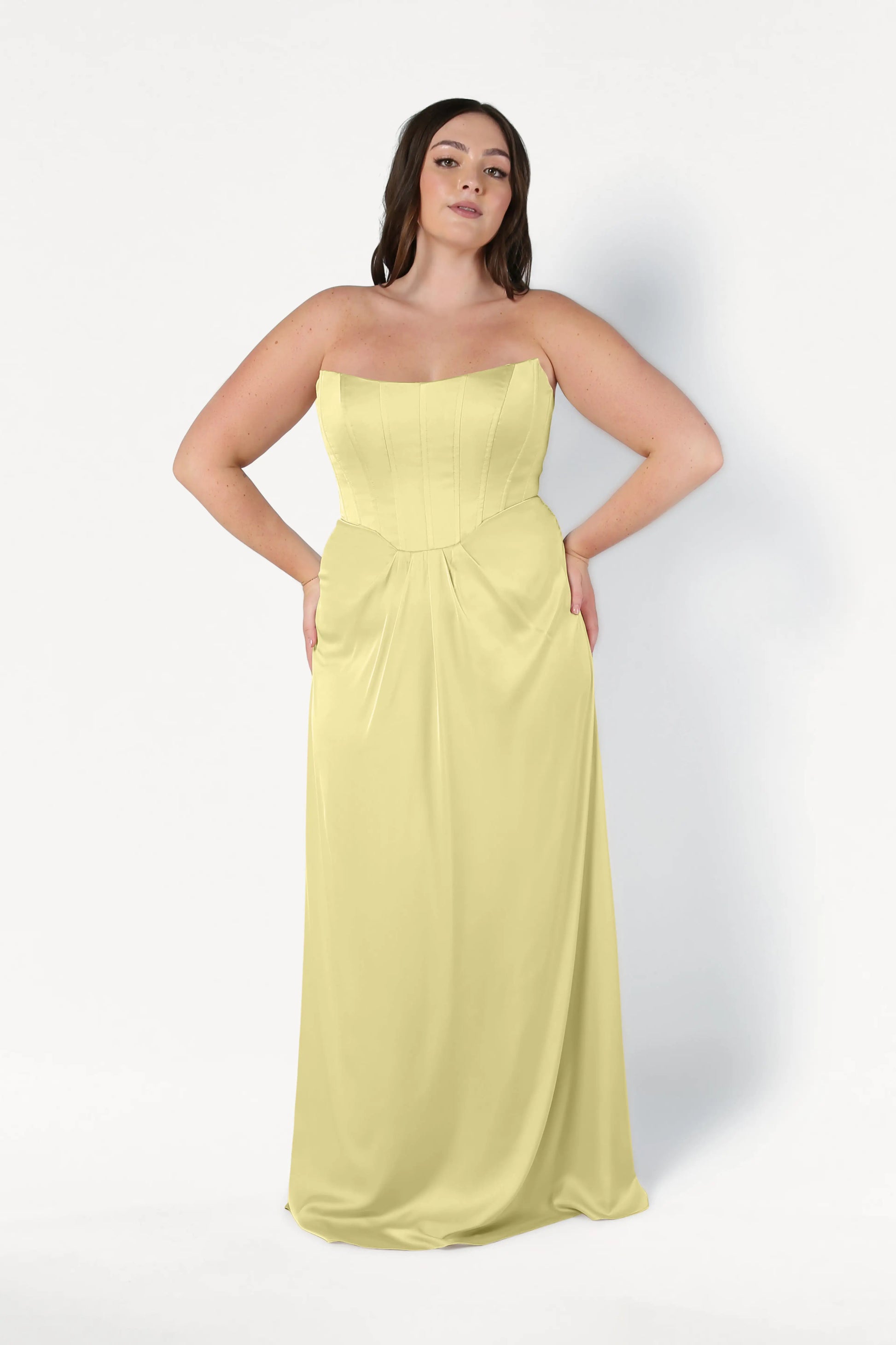 A woman poses against a plain white background, wearing the TH&TH Selena Bridesmaid Dress in Limoncello Satin—a strapless, floor-length gown with a corset bodice. Her hands rest on her hips as she faces forward.