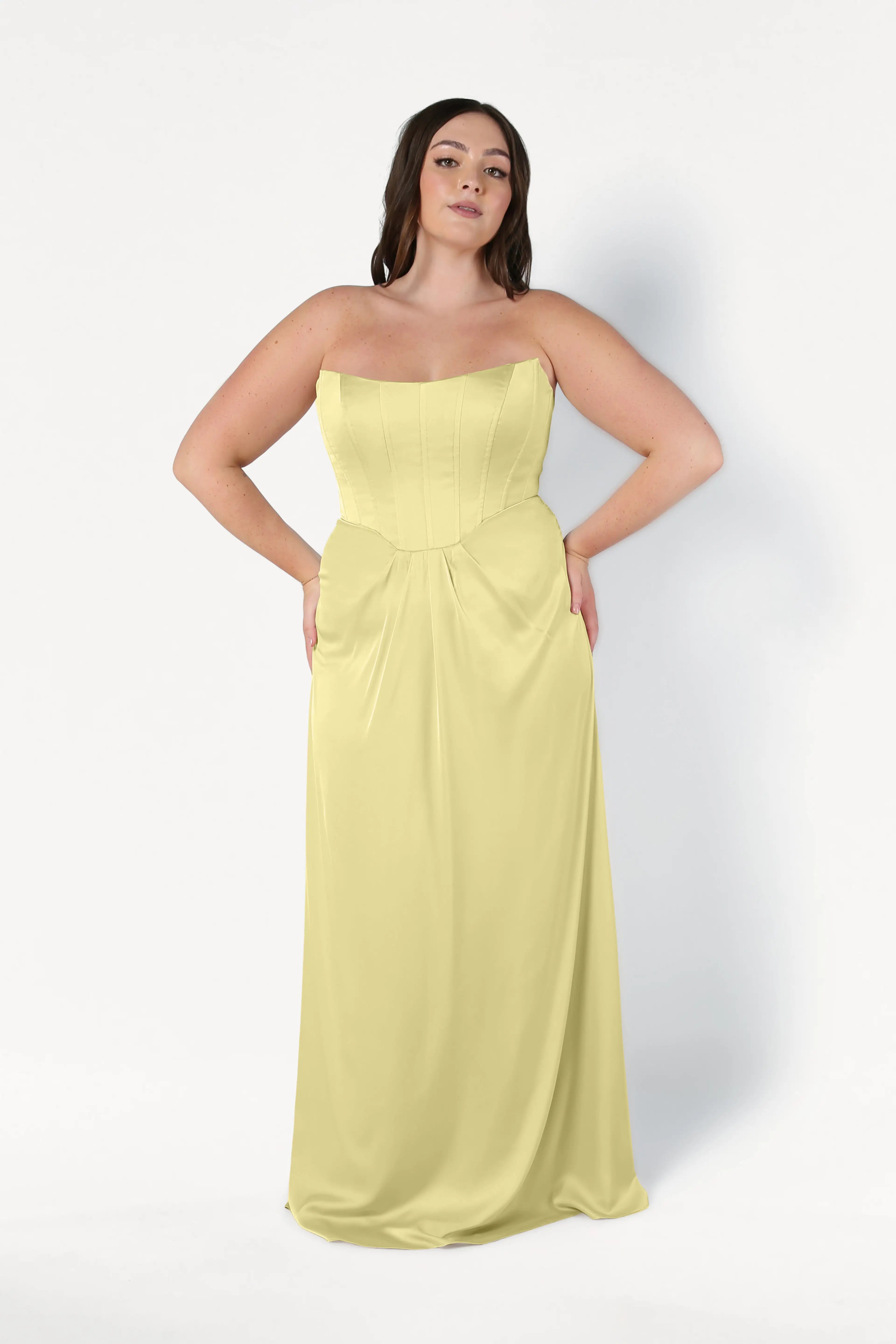 A woman poses against a plain white background, wearing the TH&TH Selena Bridesmaid Dress in Limoncello Satin—a strapless, floor-length gown with a corset bodice. Her hands rest on her hips as she faces forward.