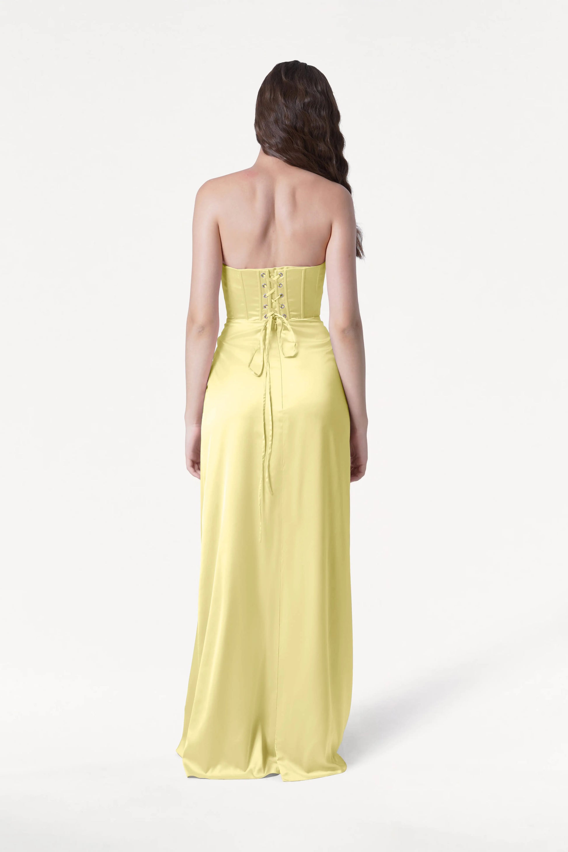 A woman with long brown hair faces away, modeling the size-inclusive Selena Bridesmaid Dress in Limoncello Satin by TH&TH—a strapless, floor-length gown with a corset bodice and lace-up back—against a plain white background.